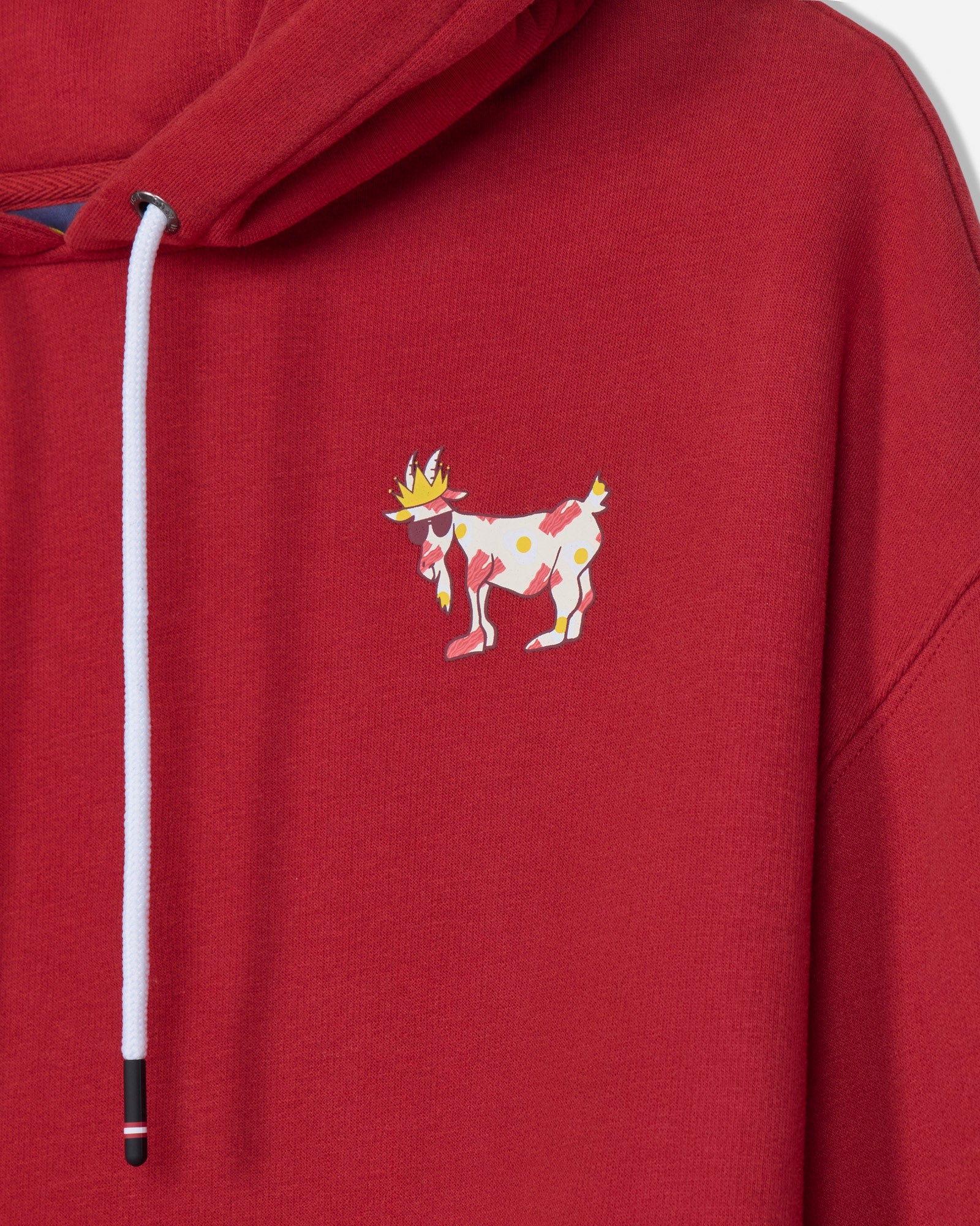 A close-up of a red hooded sweatshirt featuring a white drawstring and a small colorful embroidered goat character on the left chest.