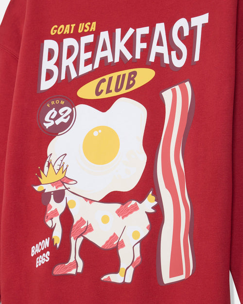 A close-up of a red graphic sweatshirt featuring a cartoon breakfast theme with the text 'GOAT USA BREAKFAST CLUB' and 'BACON EGGS'. The design includes a white goat wearing a yellow crown and sunglasses, a large fried egg, and a striped bacon slice.