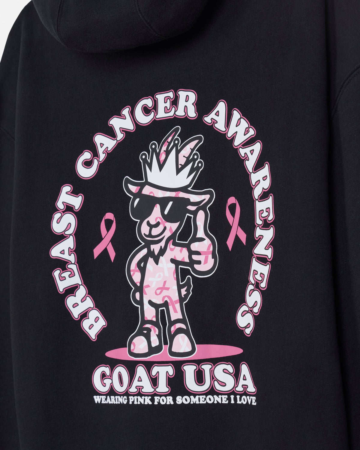 Close-up of a black hoodie featuring a large breast cancer awareness graphic on the back. The design includes a cartoon goat character wearing sunglasses and a crown, pink awareness ribbons, and the text 'BREAST CANCER AWARENESS' and 'GOAT USA'.