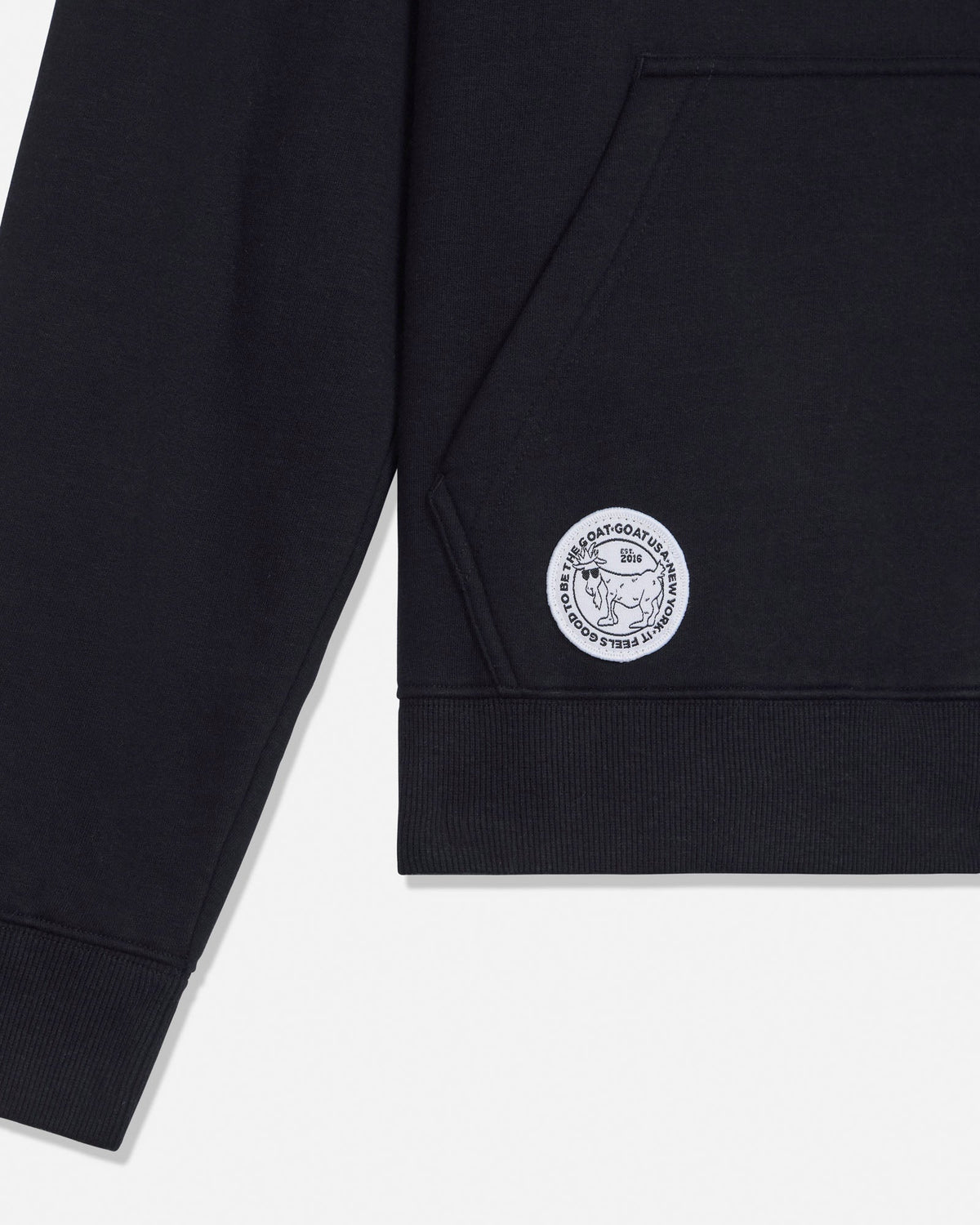 Close-up of a black sweatshirt sleeve with a circular white embroidered logo patch on the upper cuff, shown against a light gray background.