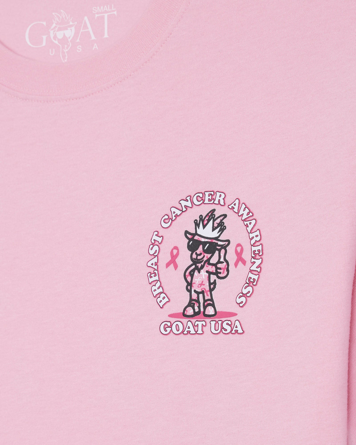 A close-up of a light pink t-shirt featuring a central graphic for Breast Cancer Awareness. The graphic shows a cartoon goat character wearing a crown and sunglasses, with pink awareness ribbons and the text 'BREAST CANCER AWARENESS' and 'GOAT USA' written around it.
