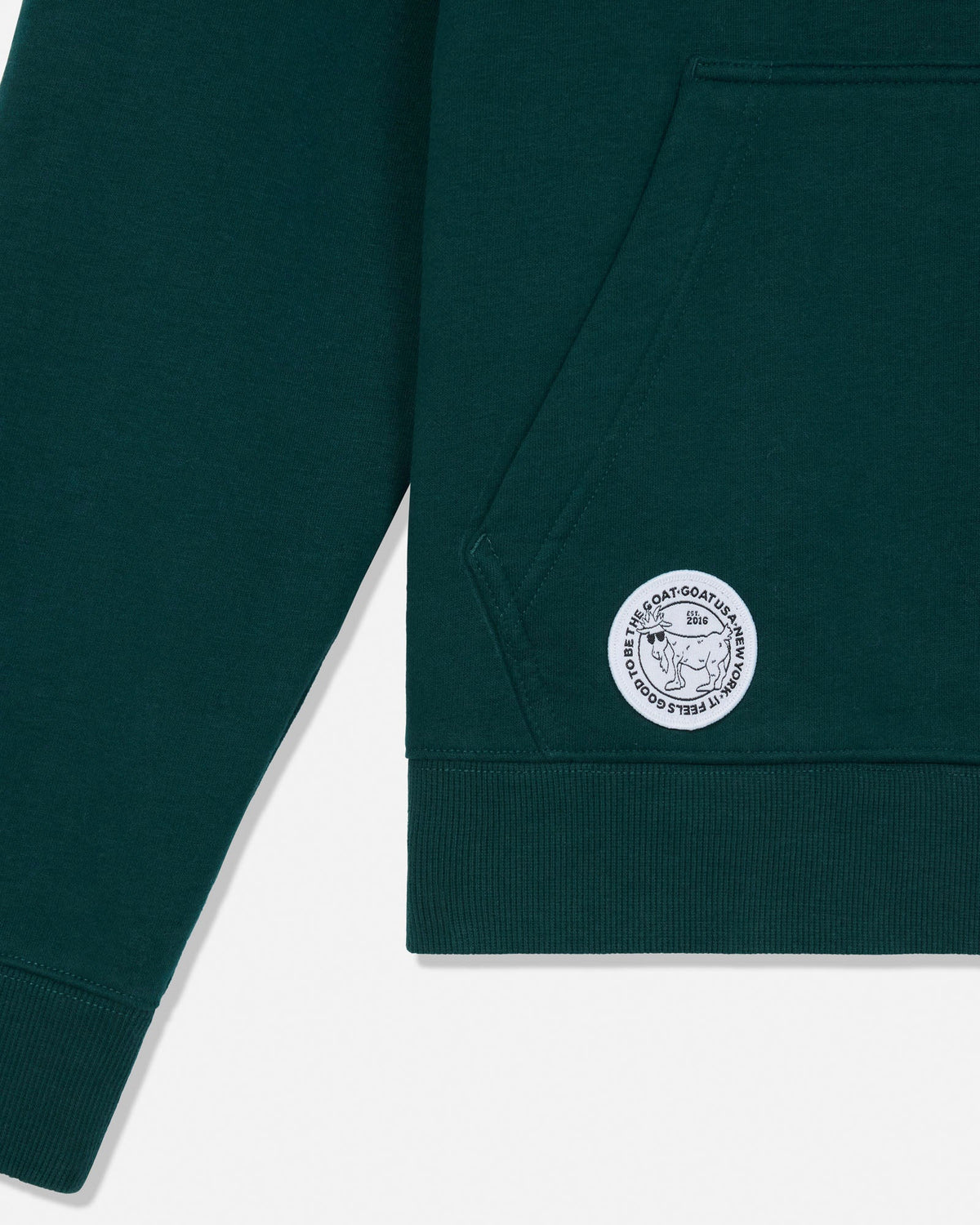 Close-up of a dark green sweatshirt sleeve and pocket area with a circular white embroidered logo patch on the pocket.