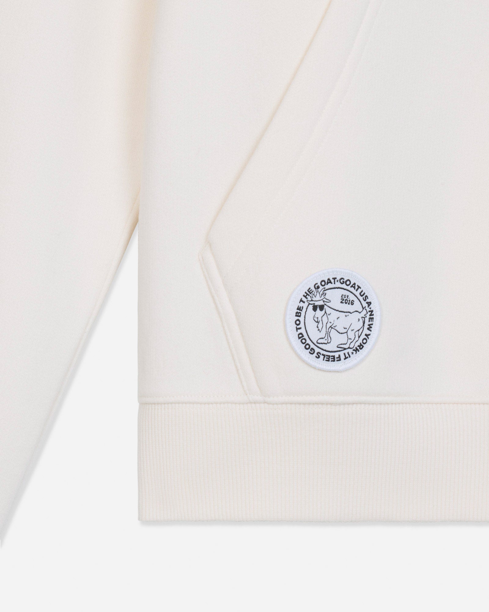 Close-up of a cream-colored sweatshirt showing the sleeve cuff, ribbed hem, and a circular embroidered patch on the chest.