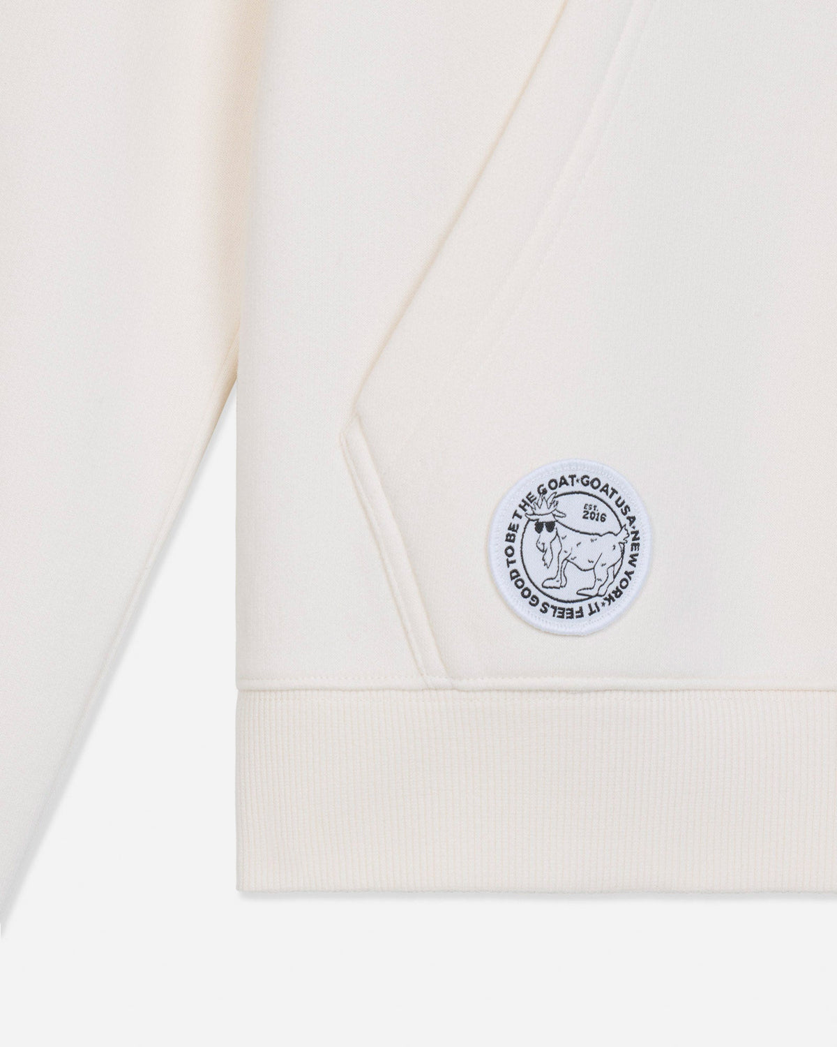 Close-up of a cream-colored sweatshirt showing the sleeve cuff, ribbed hem, and a circular embroidered patch on the chest.
