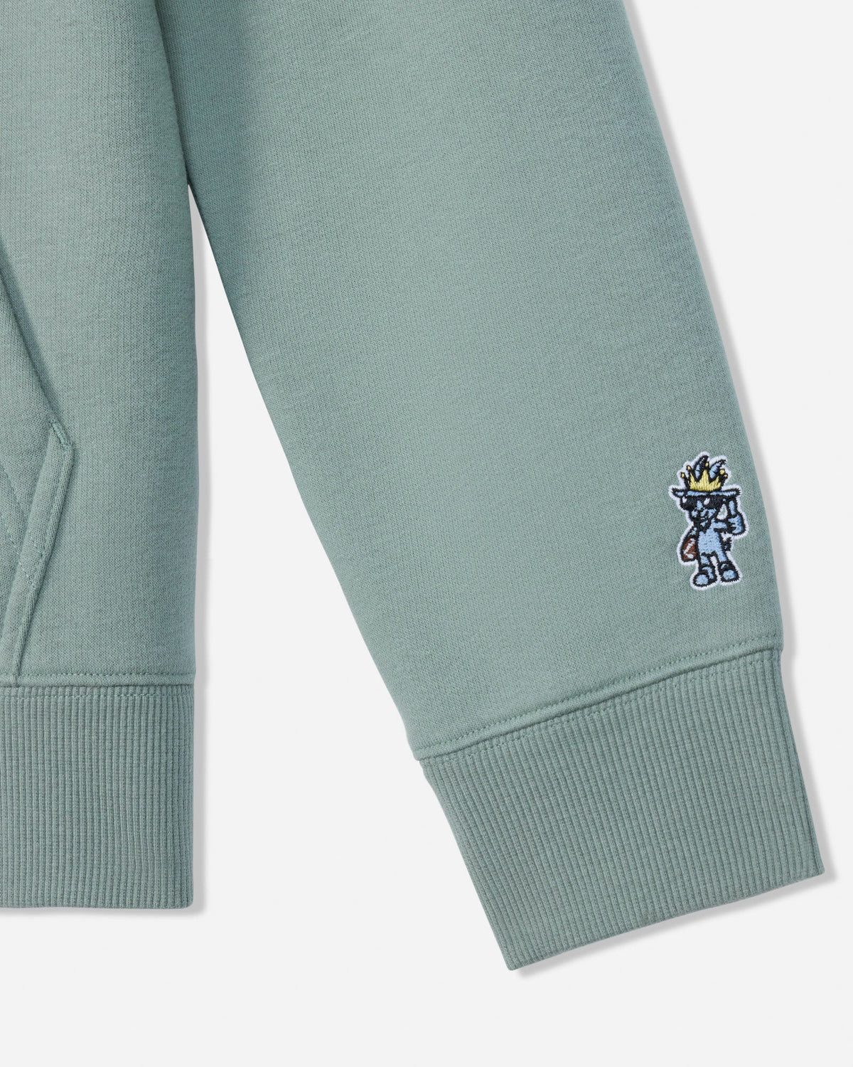 Close-up of a sage green sweatshirt sleeve with a ribbed cuff, featuring a small embroidered cartoon character on the cuff.