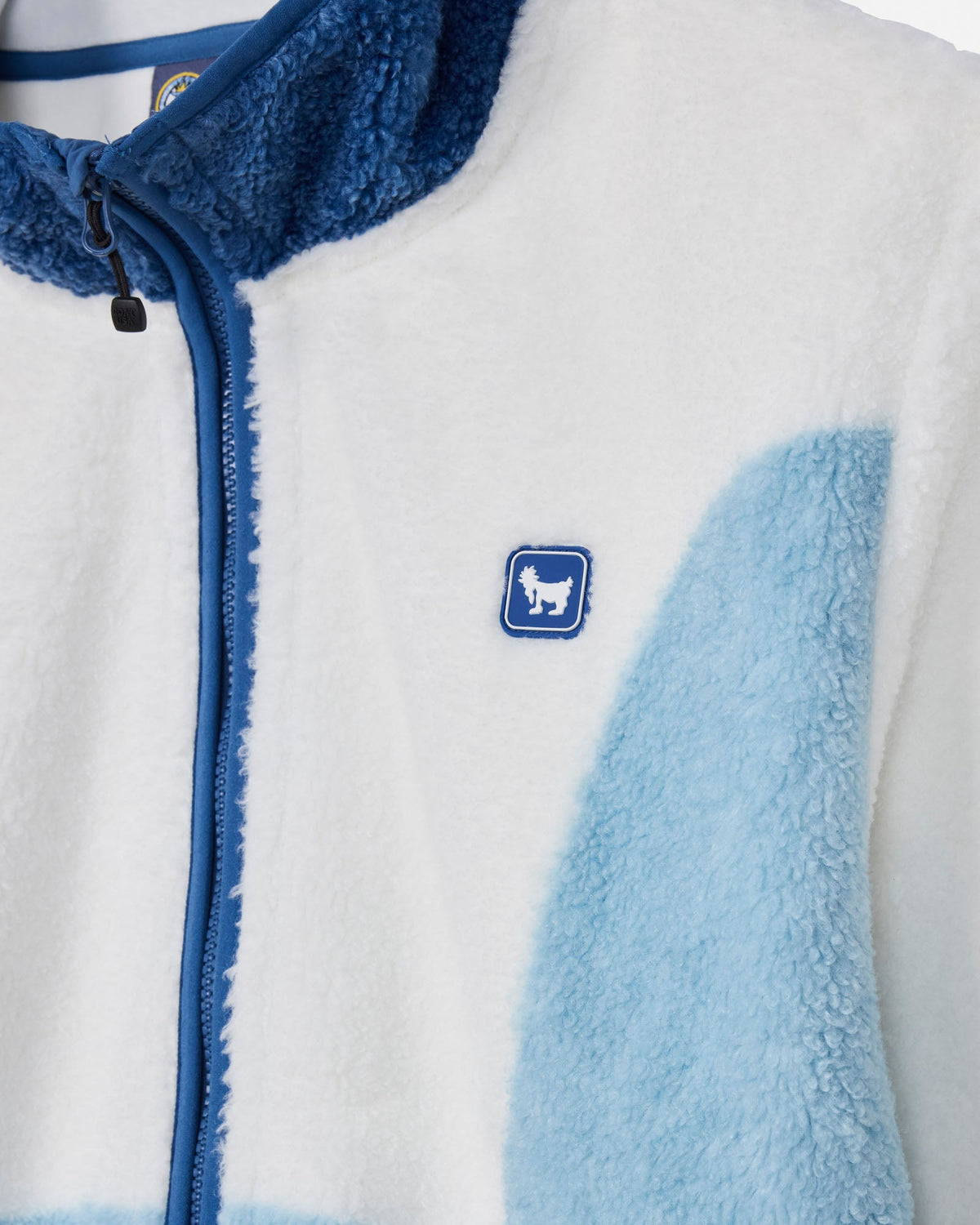 Close-up of a fleece zip jacket with a white outer shell, blue collar and zipper trim, and a light blue patch on the right side. A small blue square logo with a white goat silhouette is visible on the chest.