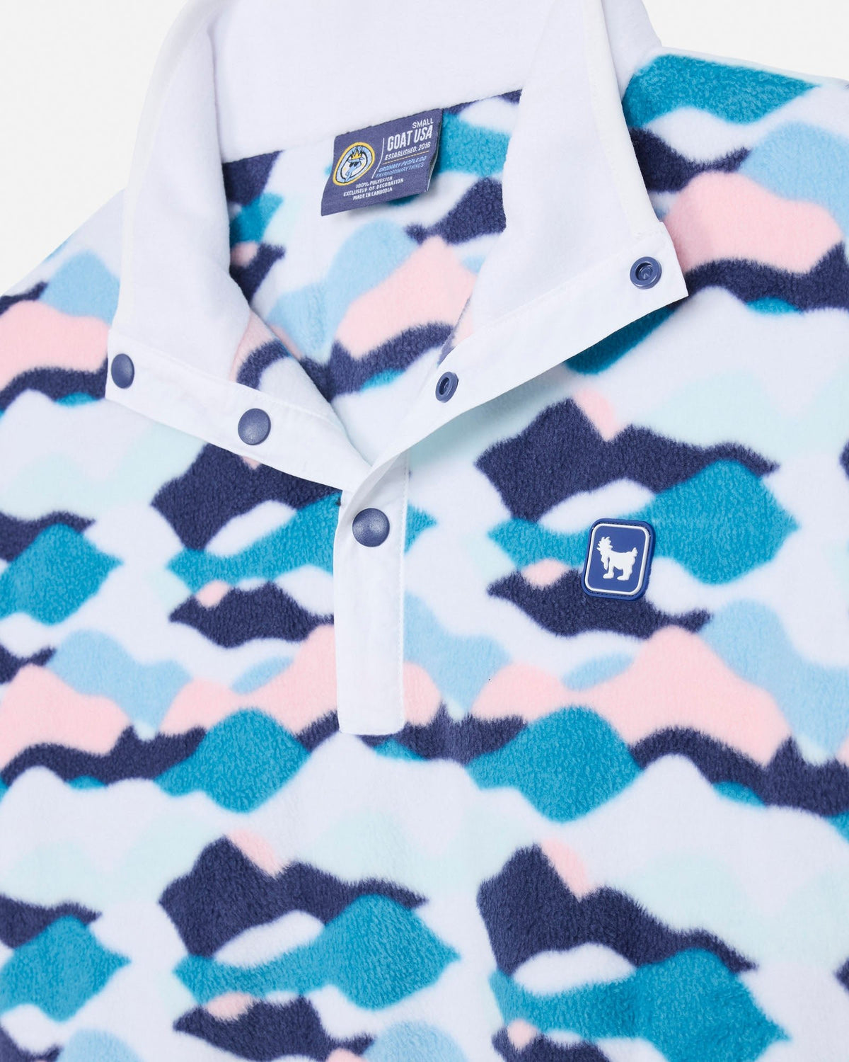 A close-up of a fleece pullover shirt with a white collar and snap-button placket, featuring a wavy abstract pattern in navy, teal, and light pink on a white background. A small blue chest patch with a white goat silhouette is visible.