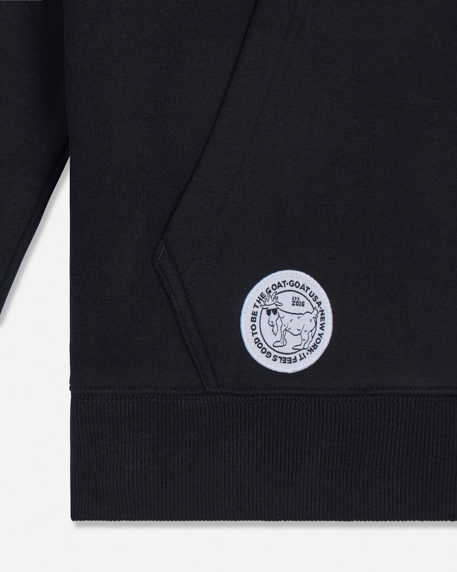 Close-up of a black ribbed sweatshirt cuff with a circular white embroidered logo patch featuring a bear and the text 'IT FEELS GOOD TO BE THE GOAT'.