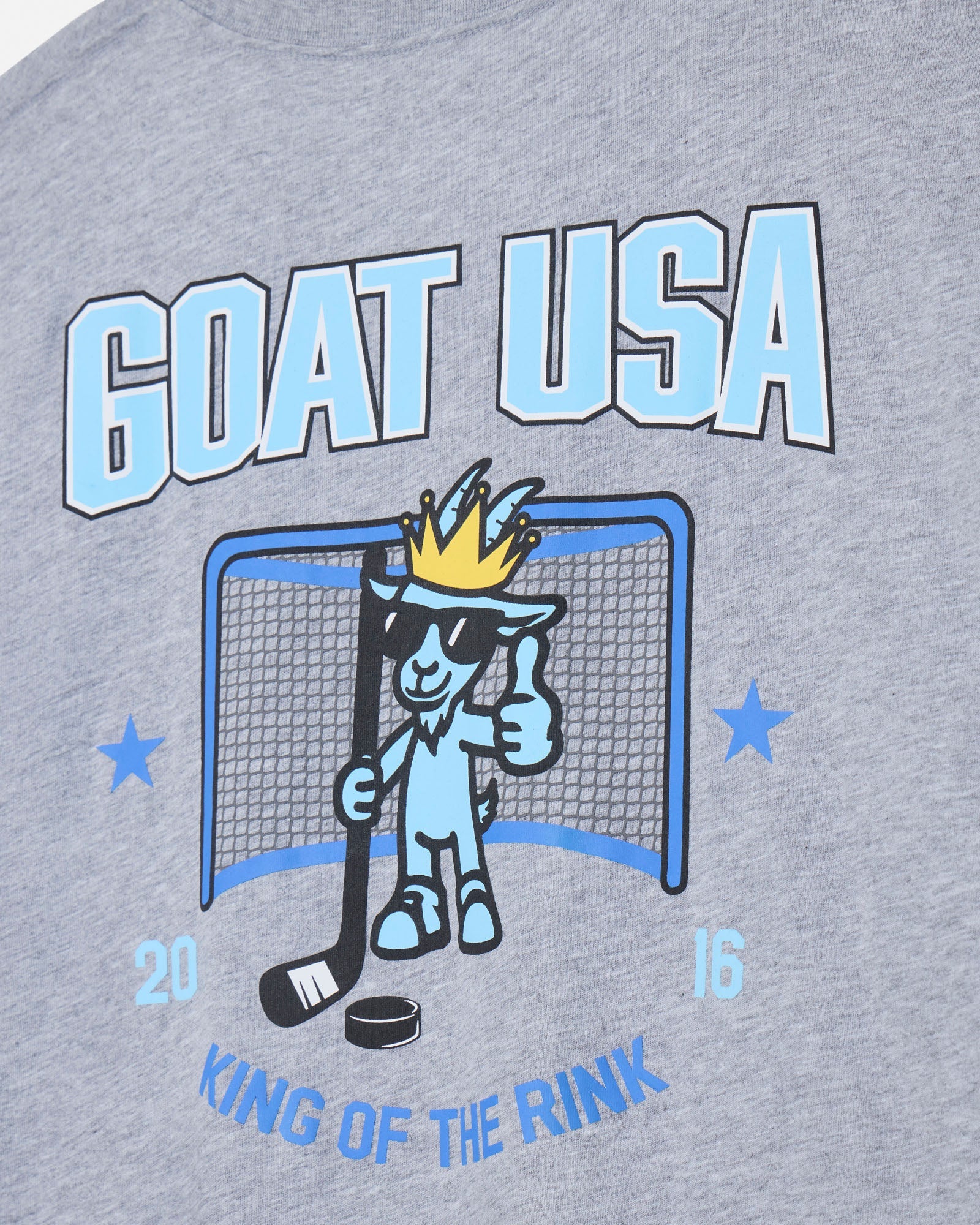 Close-up of a heather gray graphic T-shirt featuring a light blue and black 'GOAT USA' design with a cartoon goat in a sunglasses and crown, plus 'KING OF THE RINK' text.