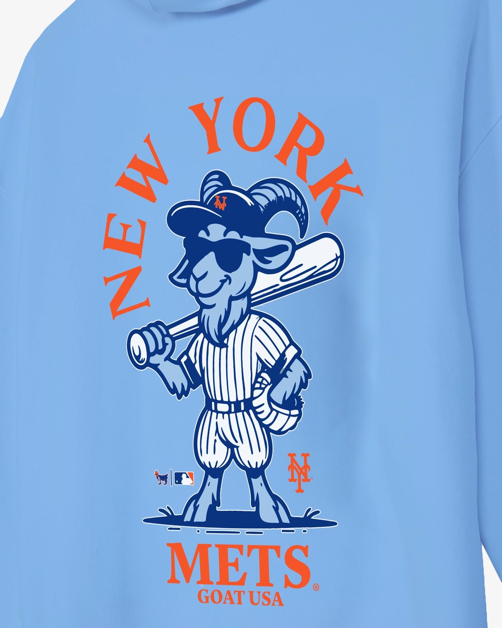 A close-up of a light blue long-sleeve shirt featuring a large New York Mets graphic print on the front. The graphic shows the team's mascot, a goat in a pinstripe baseball uniform holding a bat, with 'NEW YORK' arched above and 'METS GOAT USA' printed below in orange and navy blue.