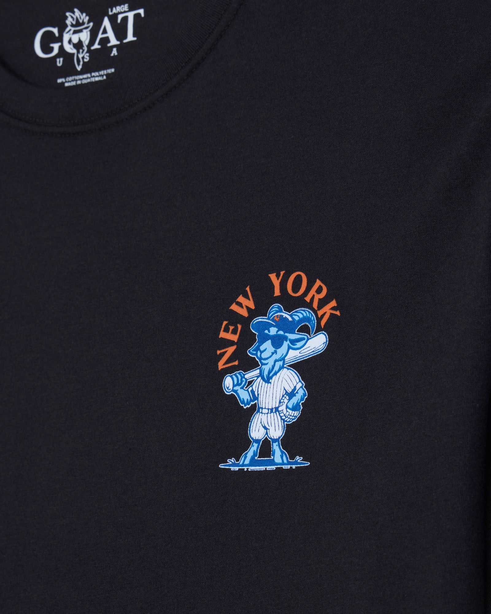 Close-up of a black t-shirt featuring a blue cartoon goat mascot holding a baseball bat, with 'NEW YORK' printed in orange arched above it. The inner neck label shows the white 'GOAT' brand logo.