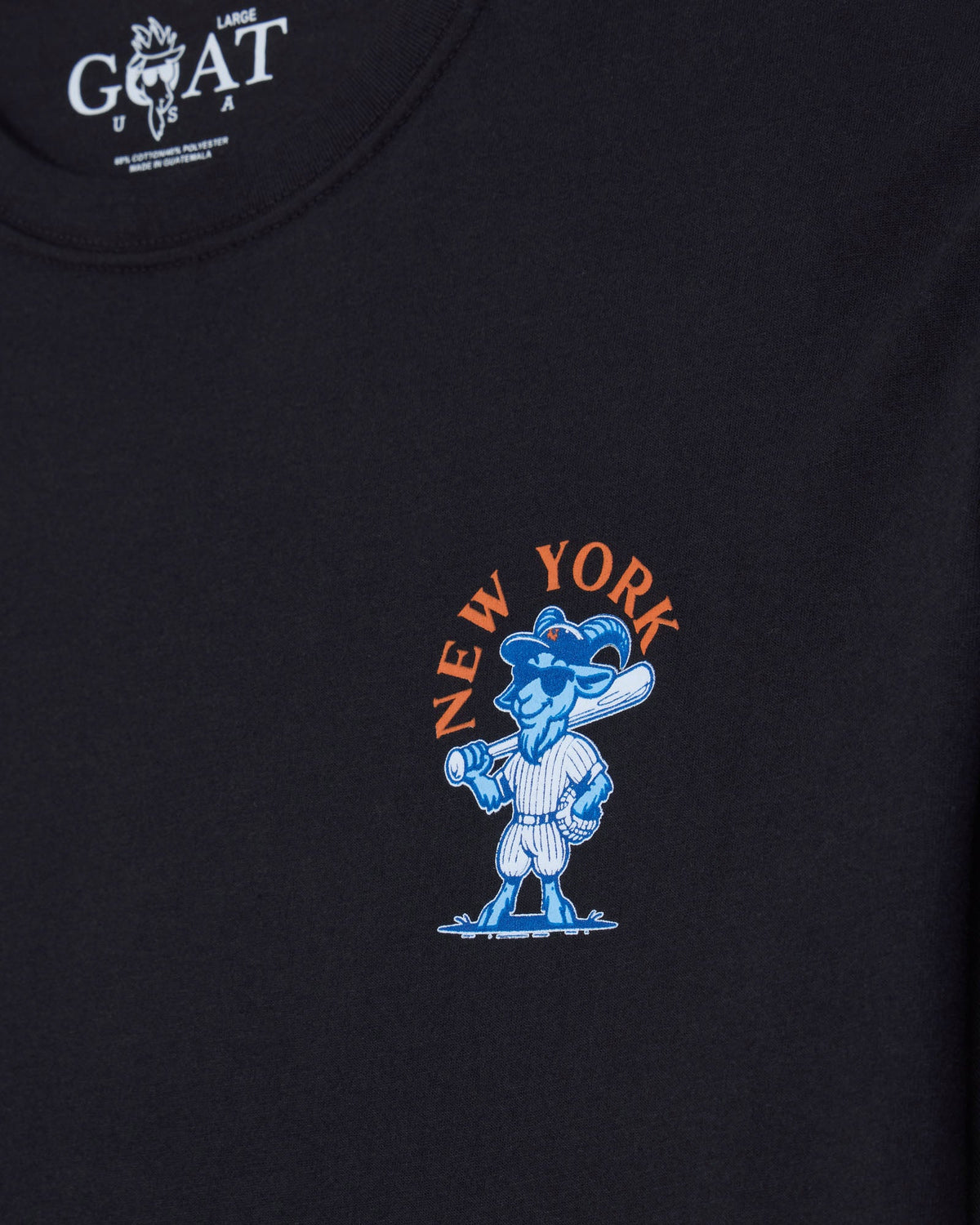 Close-up of a black t-shirt featuring a blue cartoon goat mascot holding a baseball bat, with 'NEW YORK' printed in orange arched above it. The inner neck label shows the white 'GOAT' brand logo.