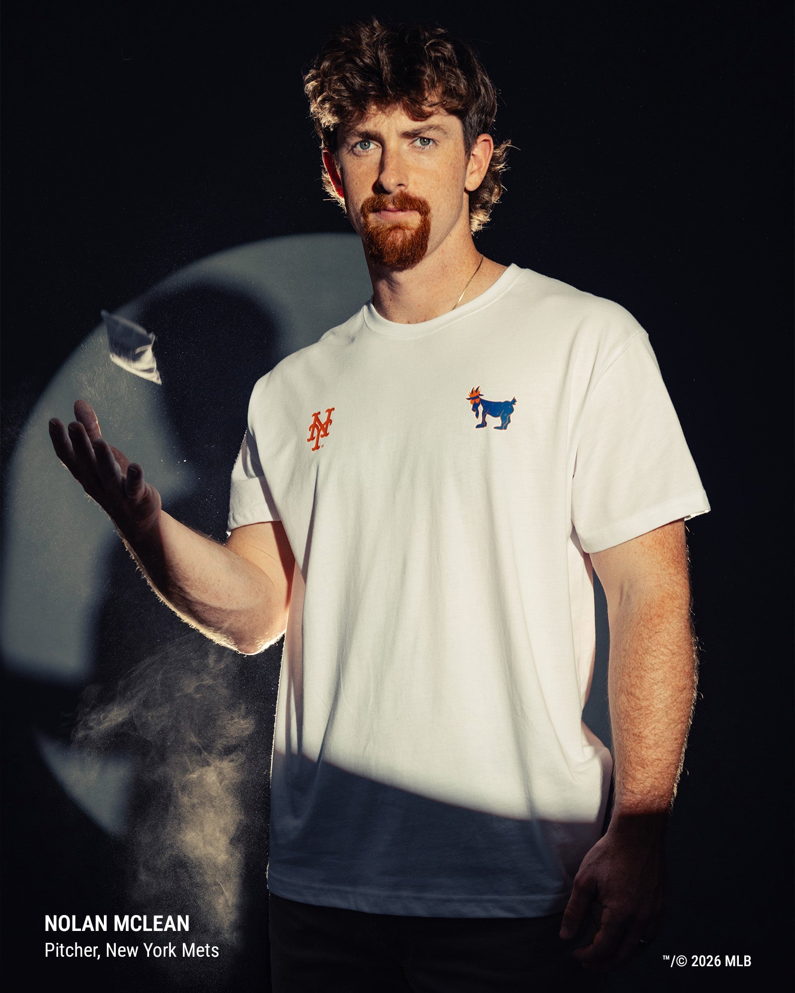 Nolan McLean wearing a white New York Mets t-shirt with a red 'NY' logo on the right chest and a blue goat logo on the left chest, posed against a dark background with dramatic lighting.
