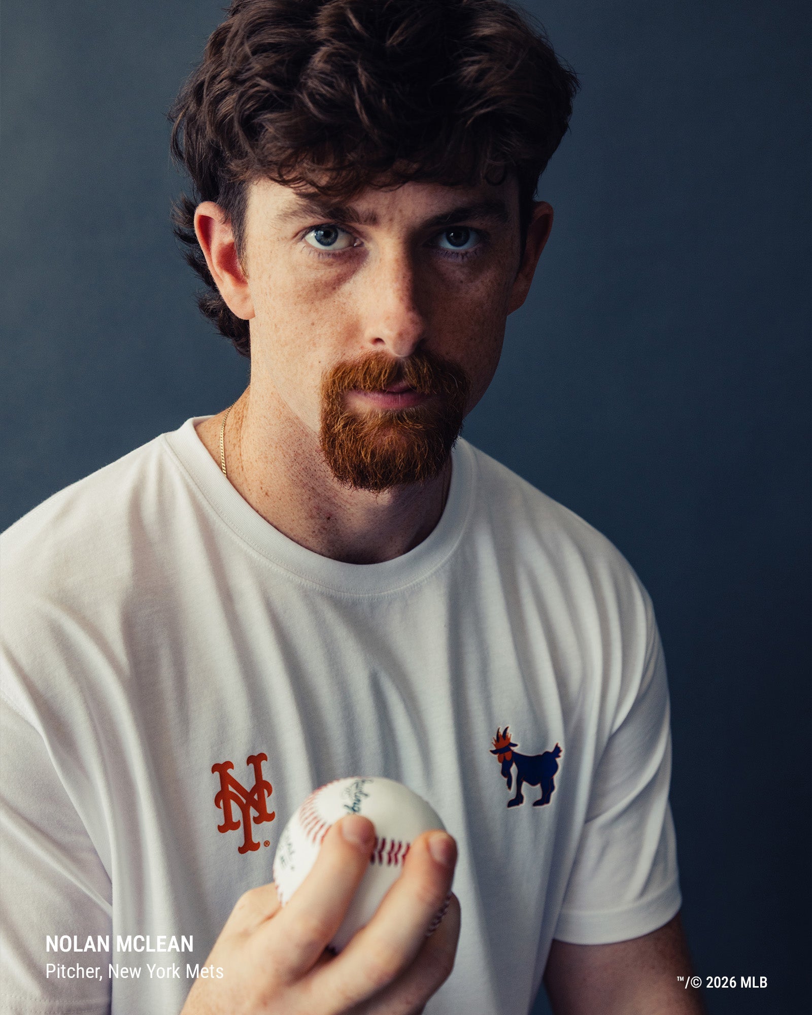 Nolan Mclean, pitcher for the New York Mets, wearing a white t-shirt with the team's red and navy goat logos, holding a baseball against a dark blue-gray background.