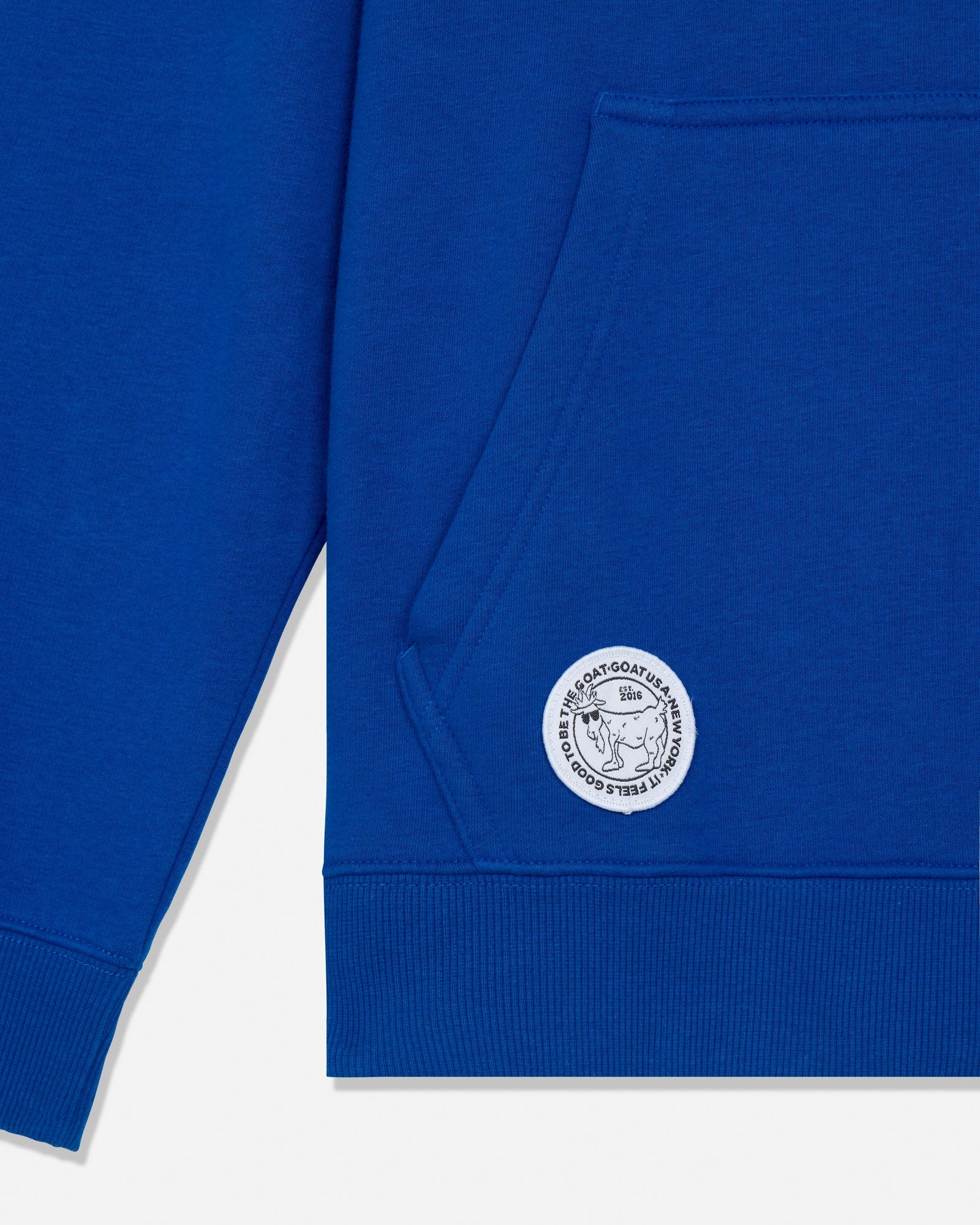Close-up of a royal blue sweatshirt showing the sleeve, ribbed hem, and a circular white embroidered logo patch on the lower front.