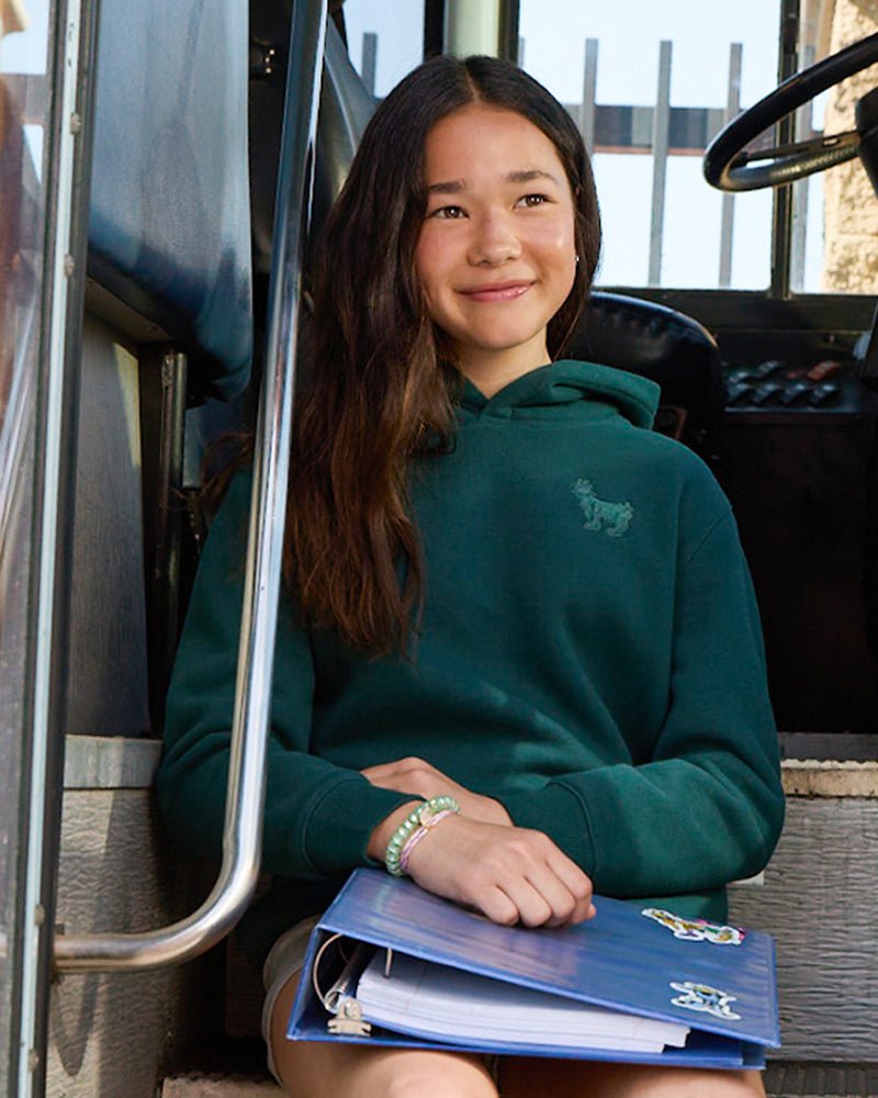 A dark teal-green pullover hoodie with a small tonal embroidered logo on the left chest, on a female youth model.#color_sage