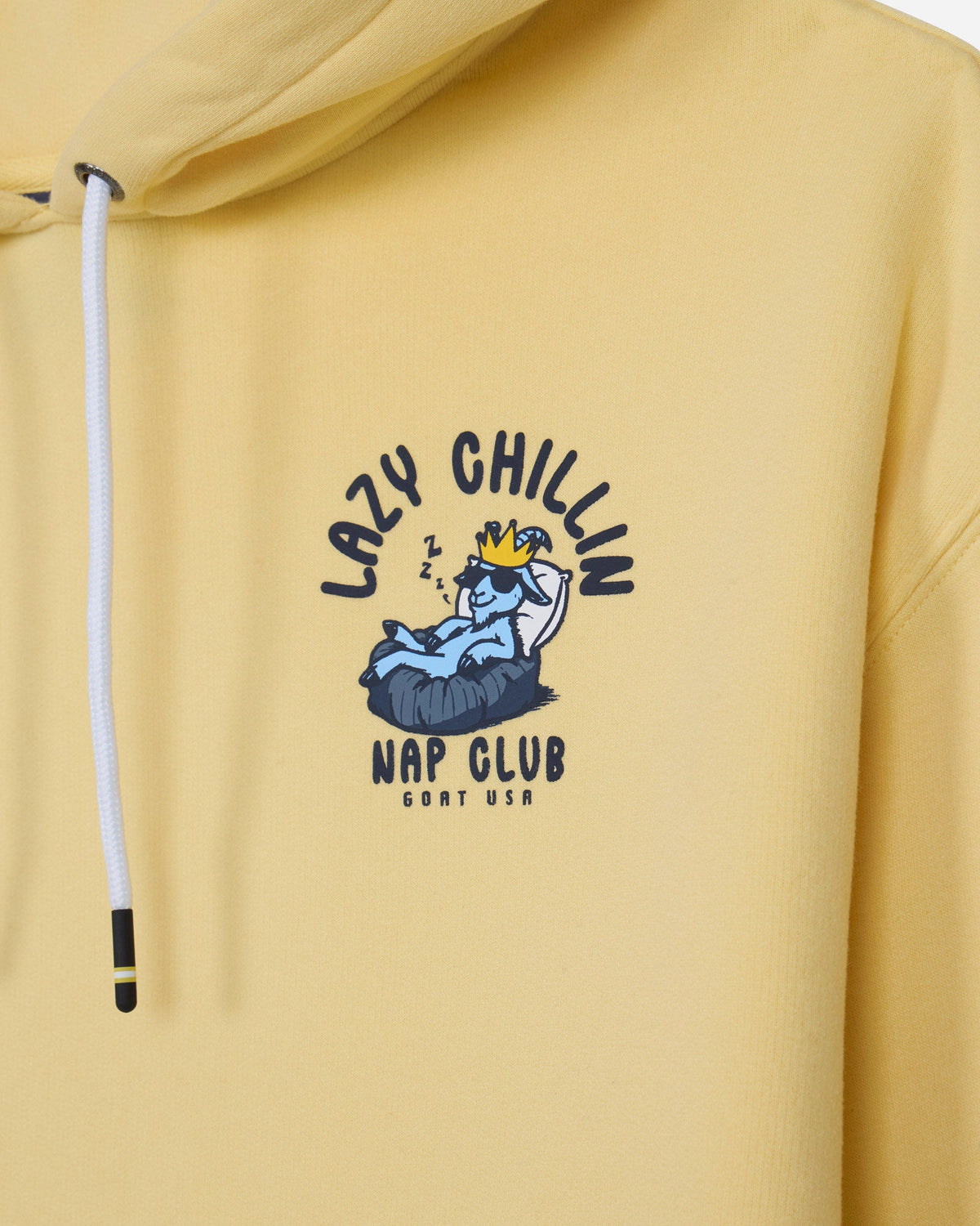 A close-up of a pale yellow hoodie featuring a central graphic print of a cartoon blue character wearing a gold crown, with the text 'LAZY CHILLIN' and 'NAP CLUB' in dark blue.