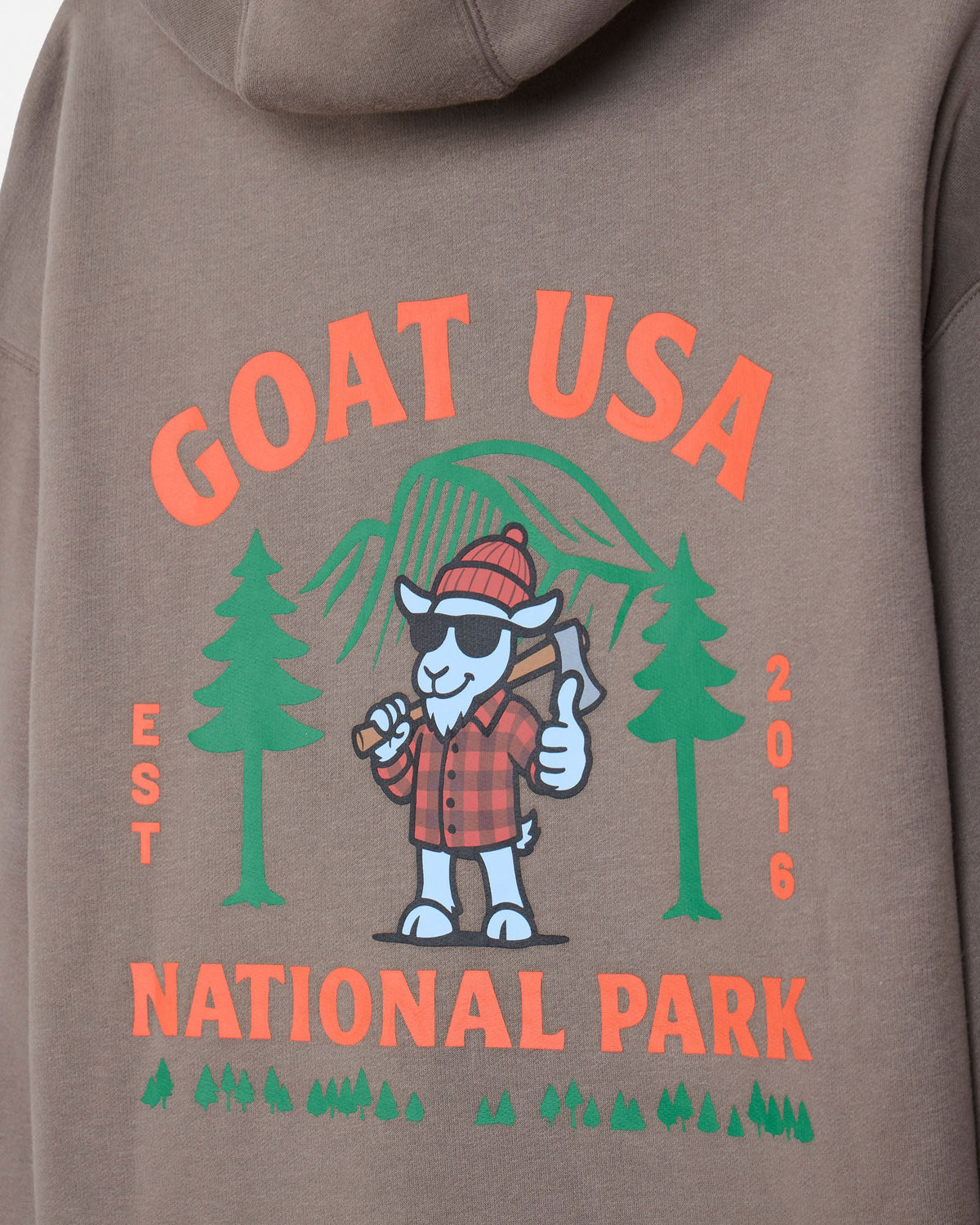 Close-up of the back of a taupe hoodie featuring a colorful graphic print with the text 'GOAT USA NATIONAL PARK', green pine trees, a cartoon goat in a red plaid shirt holding an axe, and 'EST 2016' printed vertically.