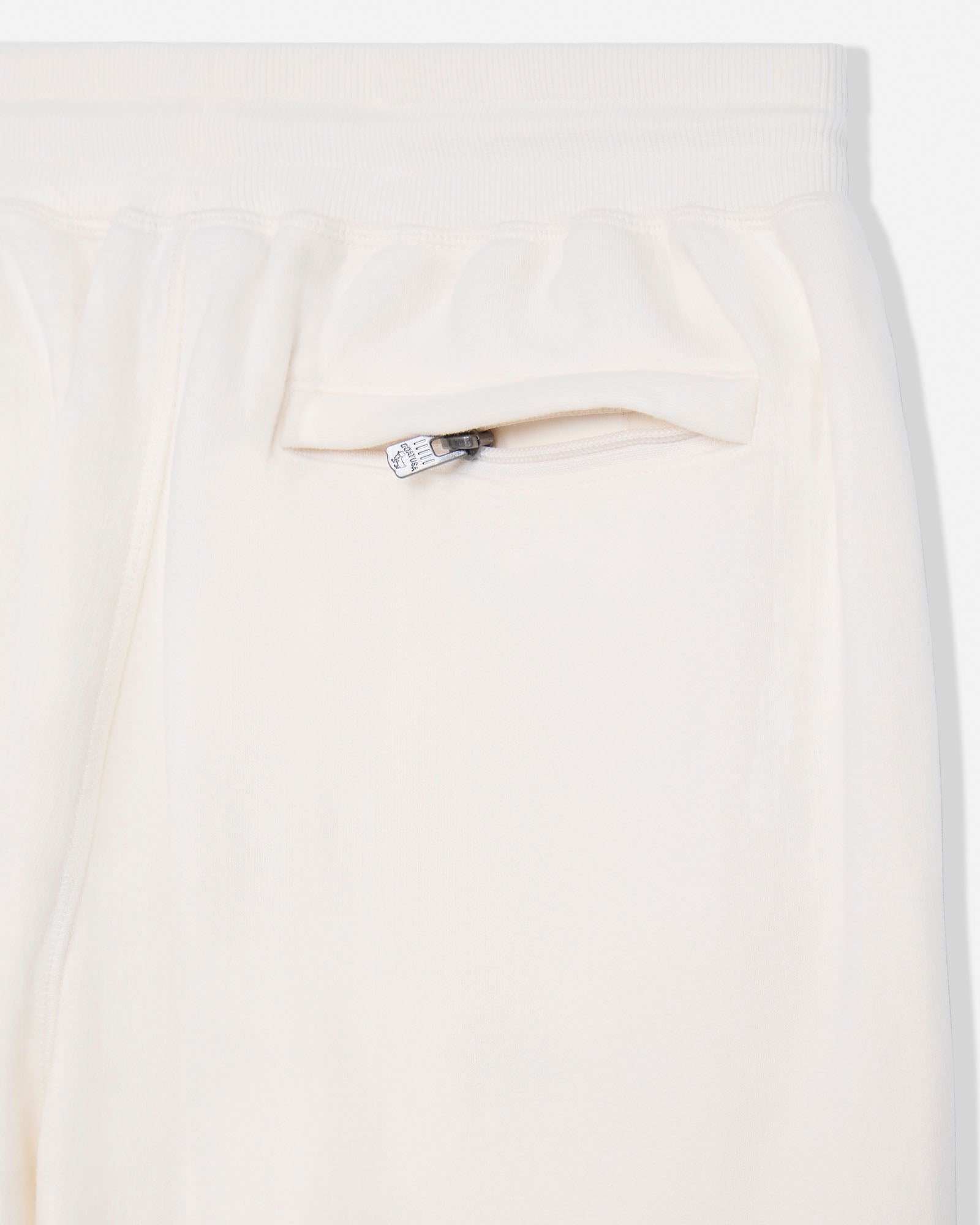Close-up of a cream-colored garment, likely a skirt or dress, featuring a ruffled waistband and a side pocket with a silver-tone zipper.#color_bone