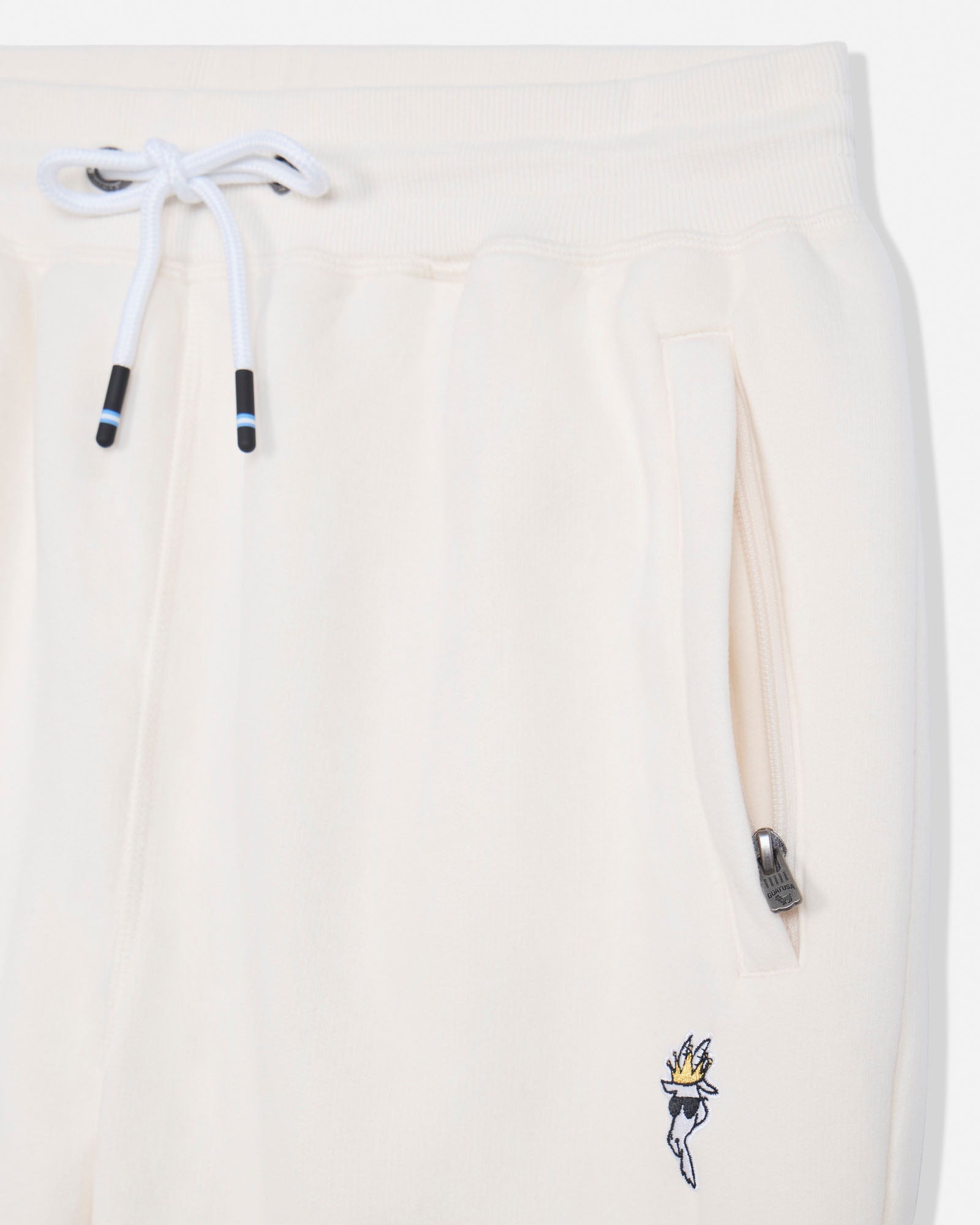 Close-up of cream-colored sweatpants featuring a white drawstring with black and blue tips, a side zippered pocket, and a small embroidered graphic near the lower hem.#color_bone