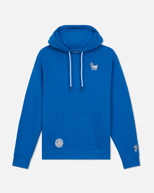 A bright blue pullover hoodie with white drawstrings, featuring a small white graphic on the left chest and a circular white graphic patch on the front pocket, displayed on a light gray background.