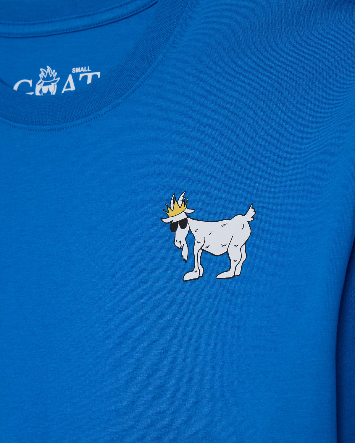 Close-up of a bright blue t-shirt featuring a white cartoon goat wearing a gold crown and black sunglasses on the chest, with a white 'SMALL GOAT' logo printed inside the collar.