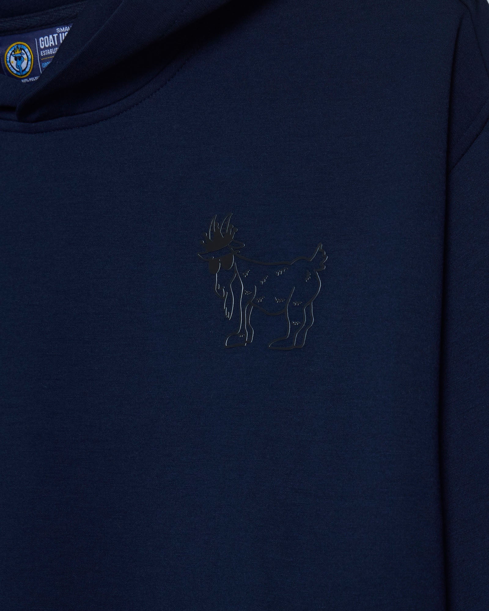 Close-up of a dark navy blue hooded sweatshirt featuring a tonal black embroidered goat logo on the left chest, with a visible GOAT USA brand tag inside the collar.#color_navy-blazer