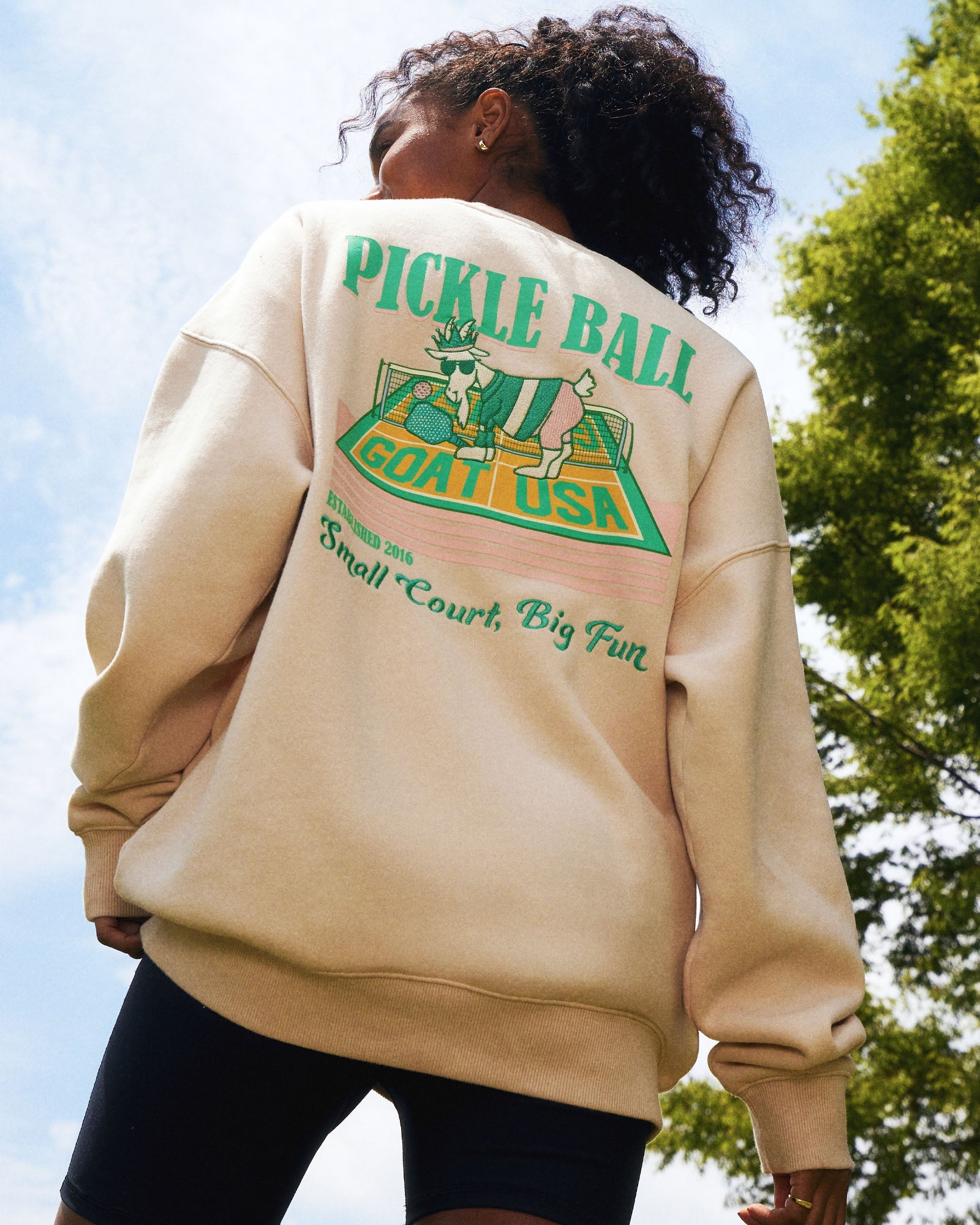 Person wearing a beige sweatshirt with 'Pickle Ball GOAT USA' graphic on the back, outdoors.