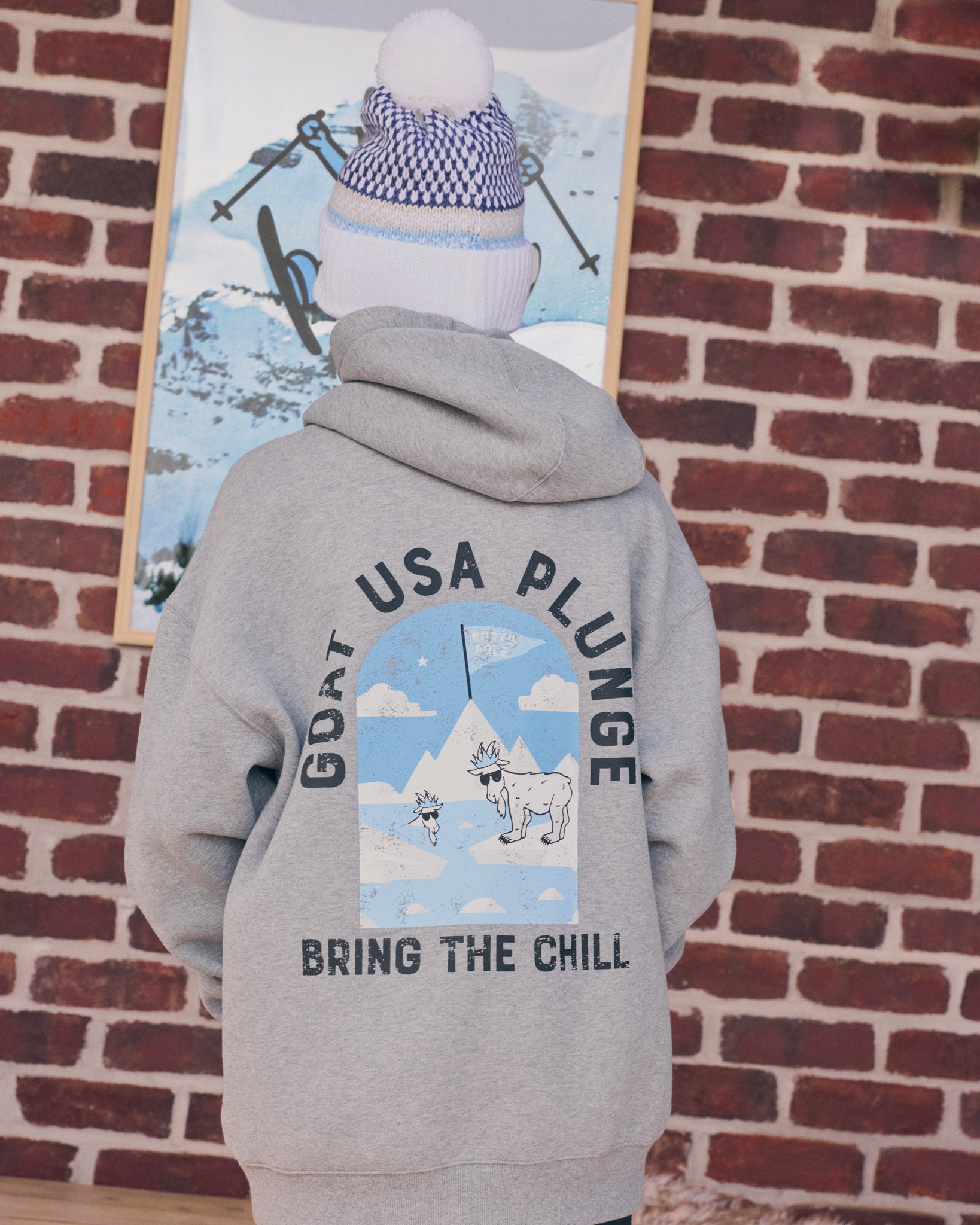 Person wearing a gray hoodie with a graphic design and text, standing against a brick wall.