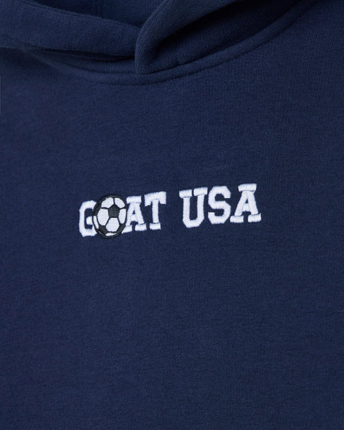 Close-up of a navy blue hoodie with 'GOAT USA' embroidered in white across the chest, where the letter O is replaced by a black-and-white soccer ball graphic.