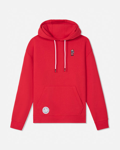A bright red pullover hoodie with white drawstrings, featuring a small embroidered character on the left chest and a circular white patch on the lower left front pocket, displayed against a light gray background.