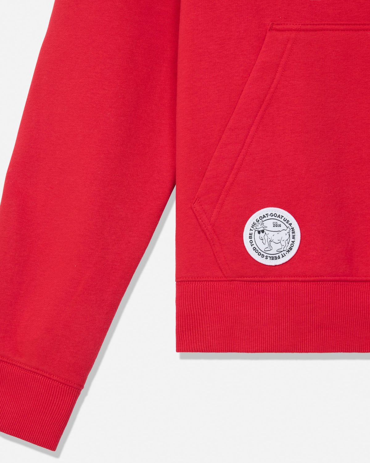 Close-up of a bright red sweatshirt showing the sleeve, ribbed cuff, front pocket, and a circular white embroidered patch on the lower front.
