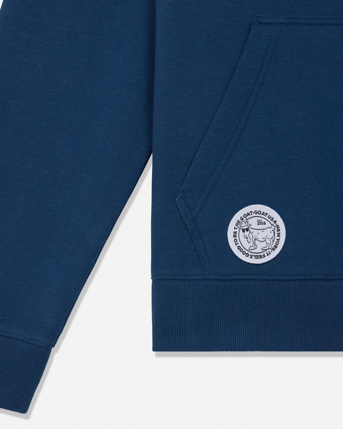 Close-up of a navy blue sweatshirt showing the sleeve, ribbed cuff, front pocket, and a circular white embroidered logo patch on the pocket.