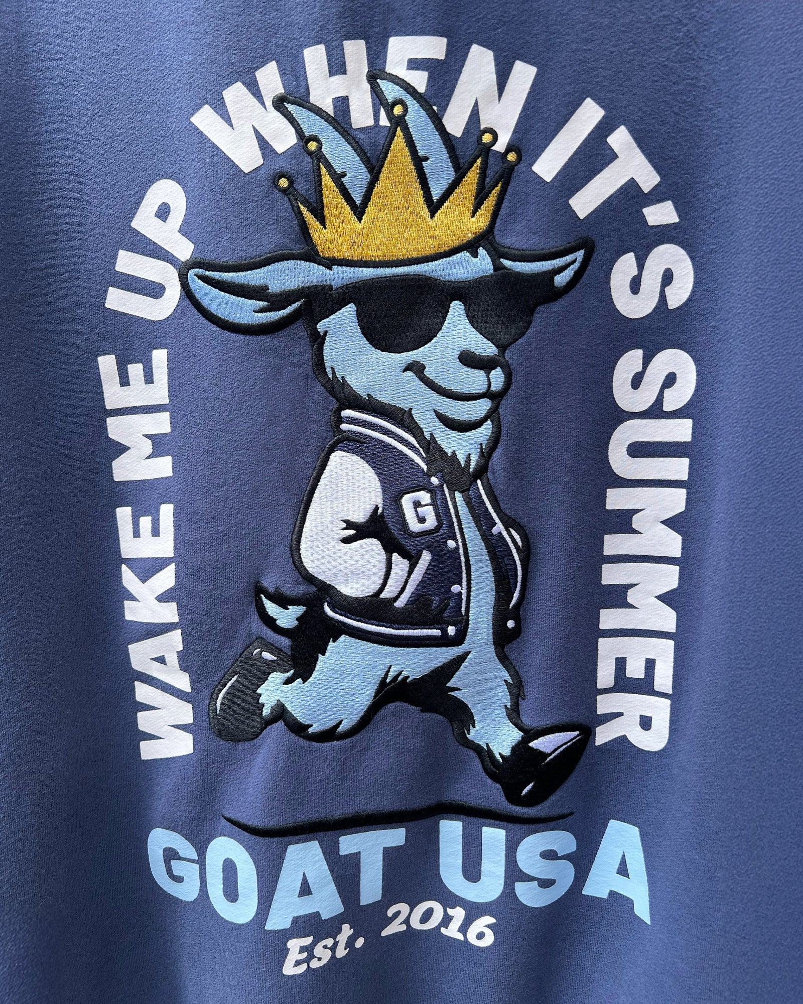 Blue crewneck with a cartoon goat wearing sunglasses and a crown, text 'Wake Me Up When It's Summer' and 'GOAT USA Est. 2016'.
