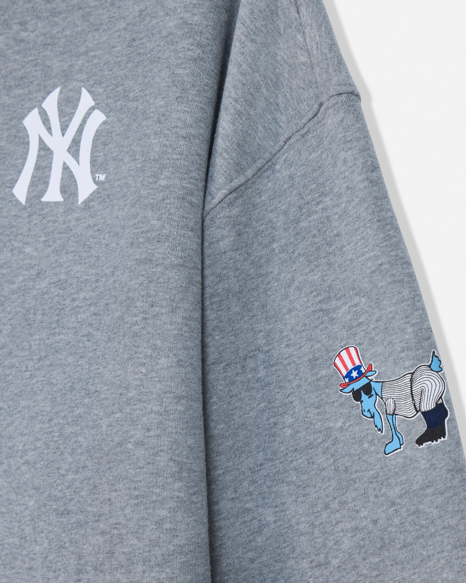 Close-up of a heather gray sweatshirt showing a white New York Yankees logo on the upper chest and a small multicolor graphic on the sleeve with a red, white, blue, and black design.