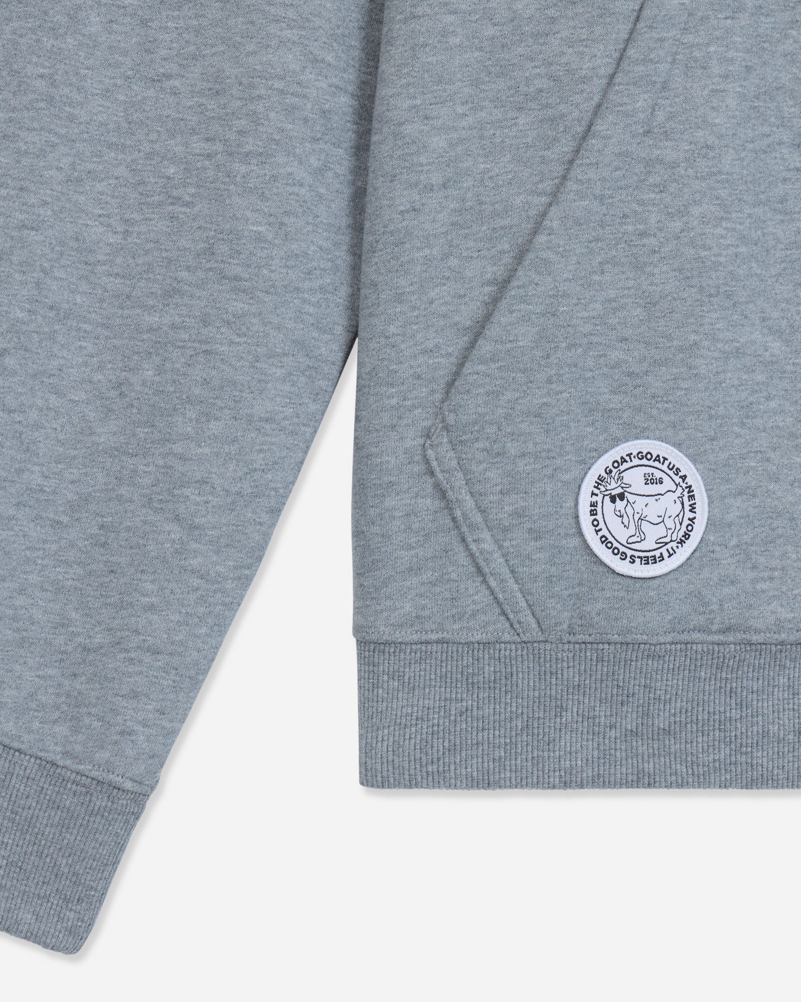 Close-up of a heather gray sweatshirt sleeve with a ribbed cuff and a circular white embroidered patch featuring a goat head and the text.#color_gray