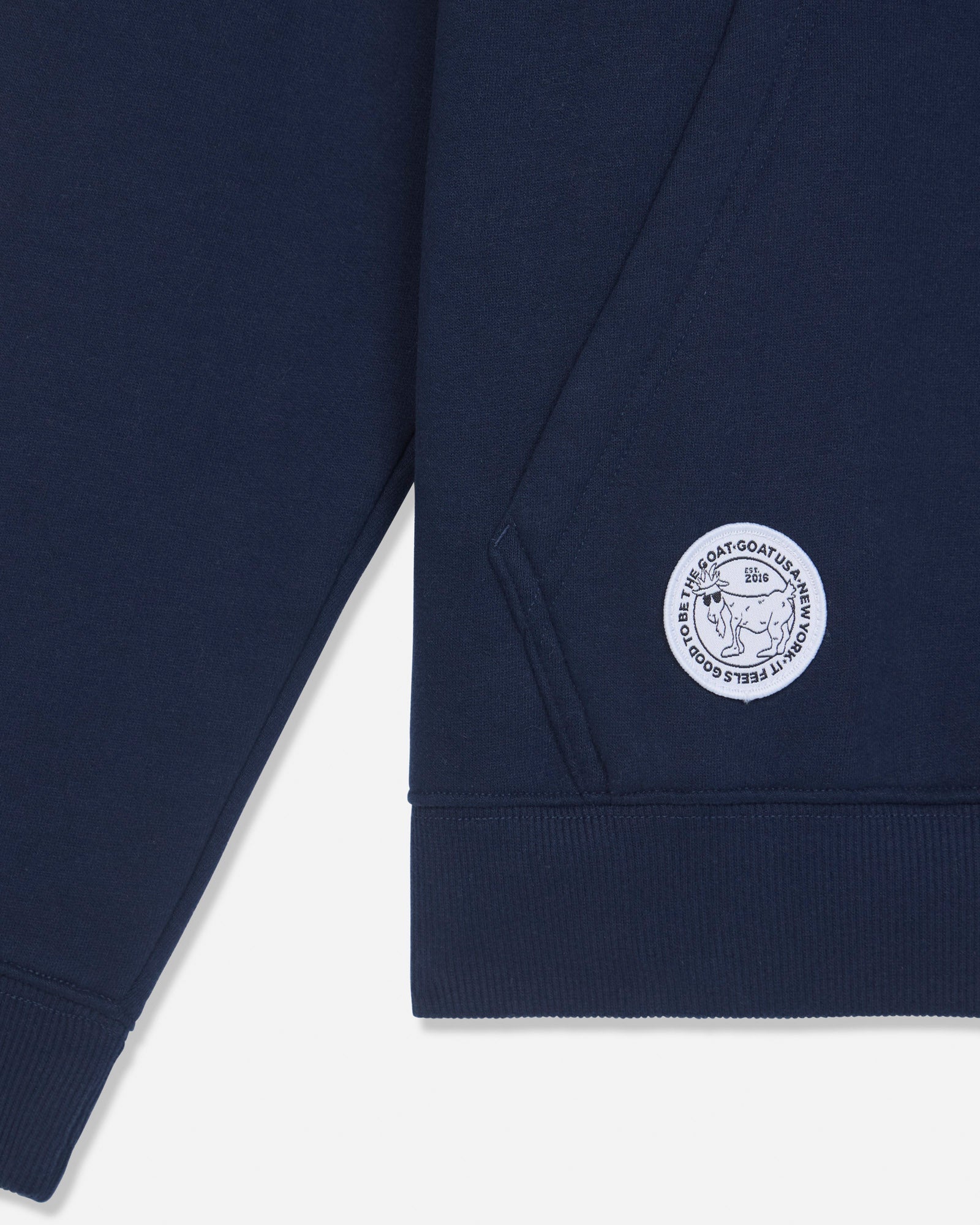 Close-up of a navy blue sweatshirt sleeve with a circular white embroidered logo patch on the cuff, shown against a light gray background.#color_navy
