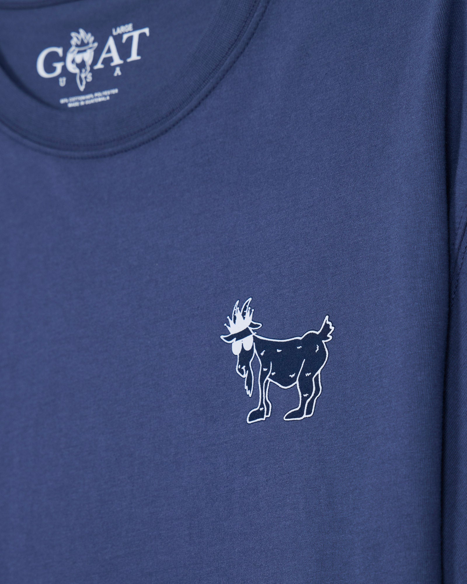 Close-up of a blue t-shirt featuring a small white line-art goat graphic on the chest and a white 'GOAT USA' logo printed inside the collar.#color_navy