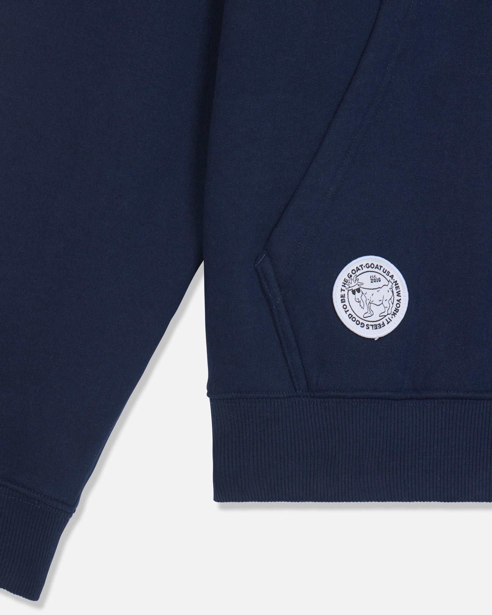Close-up of a navy blue sweatshirt sleeve showing a white circular embroidered logo patch on the cuff, photographed against a light gray background.