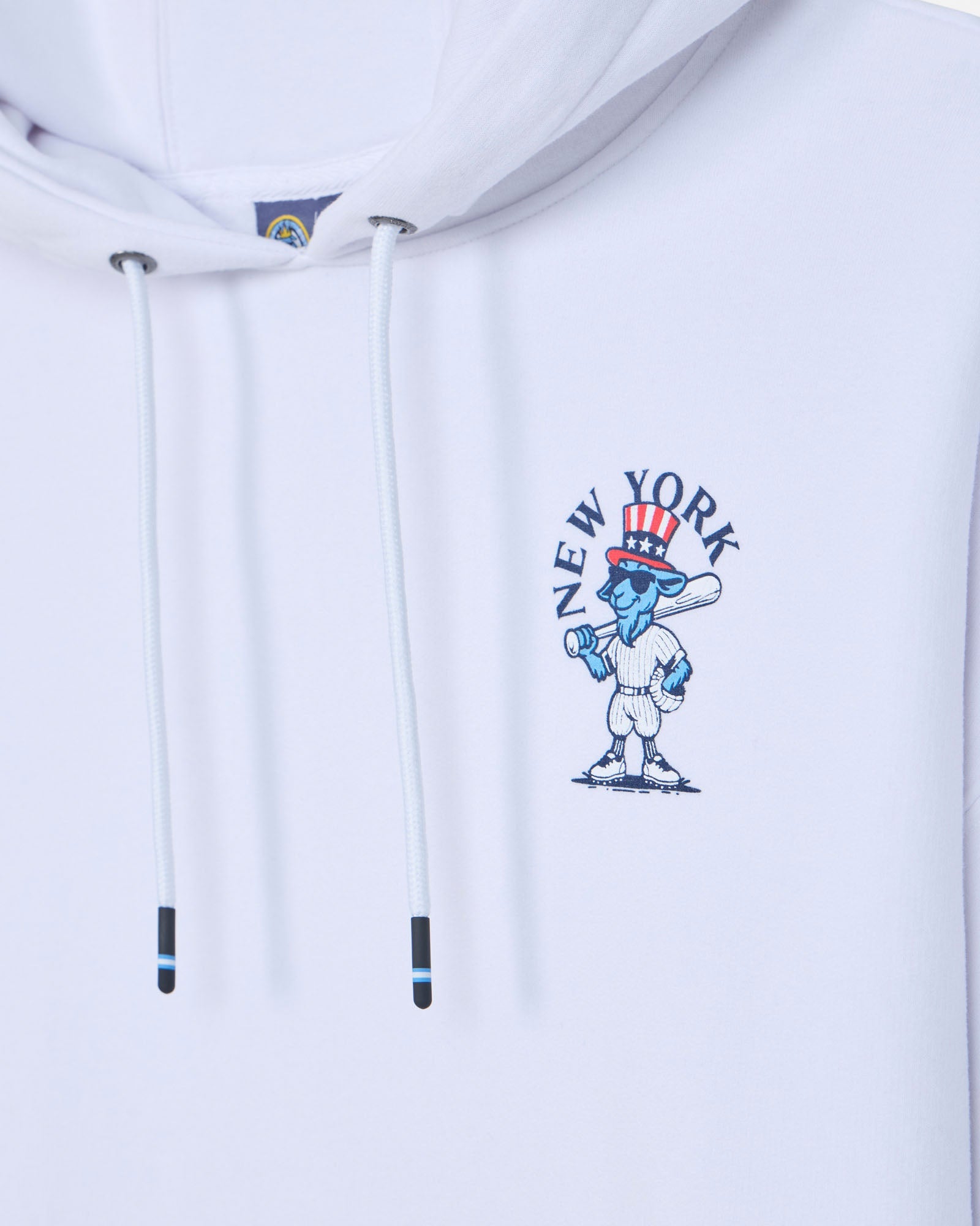 Close-up of a white pullover hoodie with white drawstrings and a blue, red, and white New York City graphic on the chest.#color_white