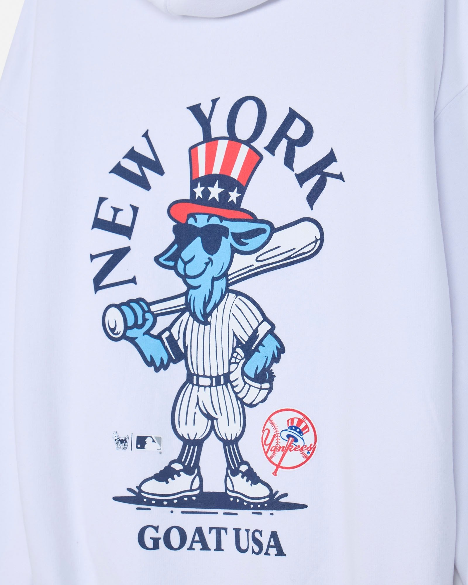 Close-up of a white long-sleeve shirt featuring a large graphic print of a blue cartoon goat baseball player with a red, white, and blue Uncle Sam hat, surrounded by the text 'NEW YORK' and 'GOAT USA'.#color_white