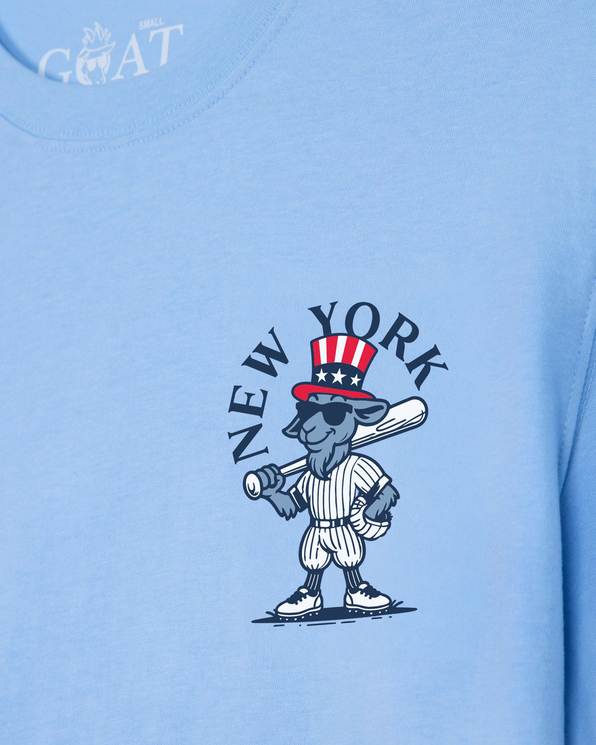 A close-up of a light blue t-shirt featuring a cartoon graphic of a gray goat baseball player wearing a red, white, and blue Uncle Sam hat, with the text 'NEW YORK' arched above him. A white 'GOAT' brand logo is visible on the inner neck collar.