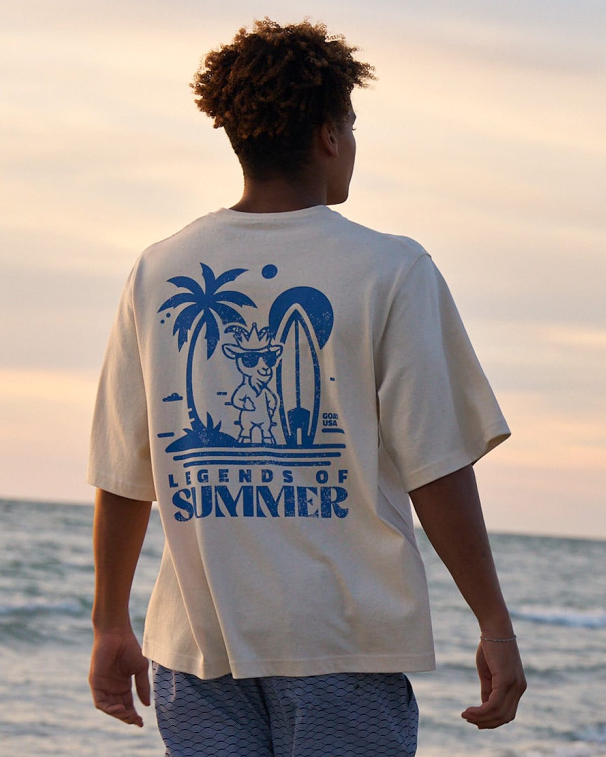 Person wearing a 'Legends of Summer' t-shirt with a beach graphic, standing by the ocean at sunset.