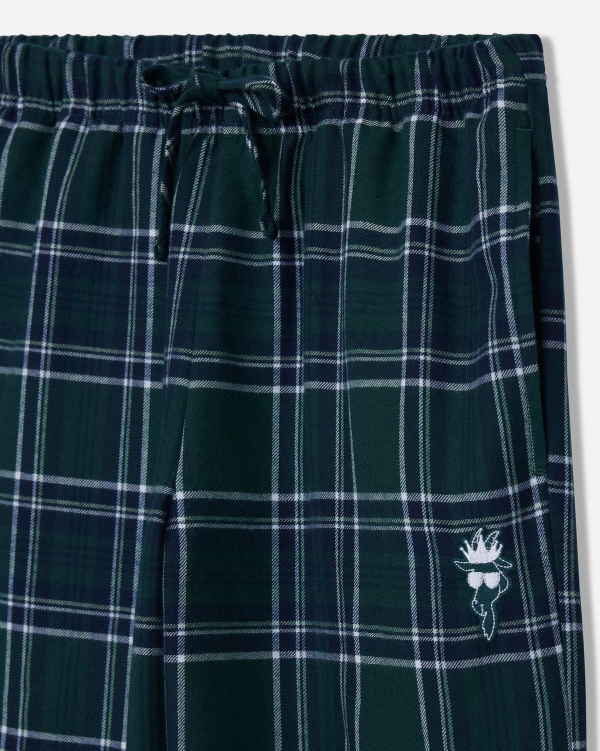 Close-up of dark green plaid lounge pants with a white drawstring and a small white embroidered goat logo near the lower right leg.