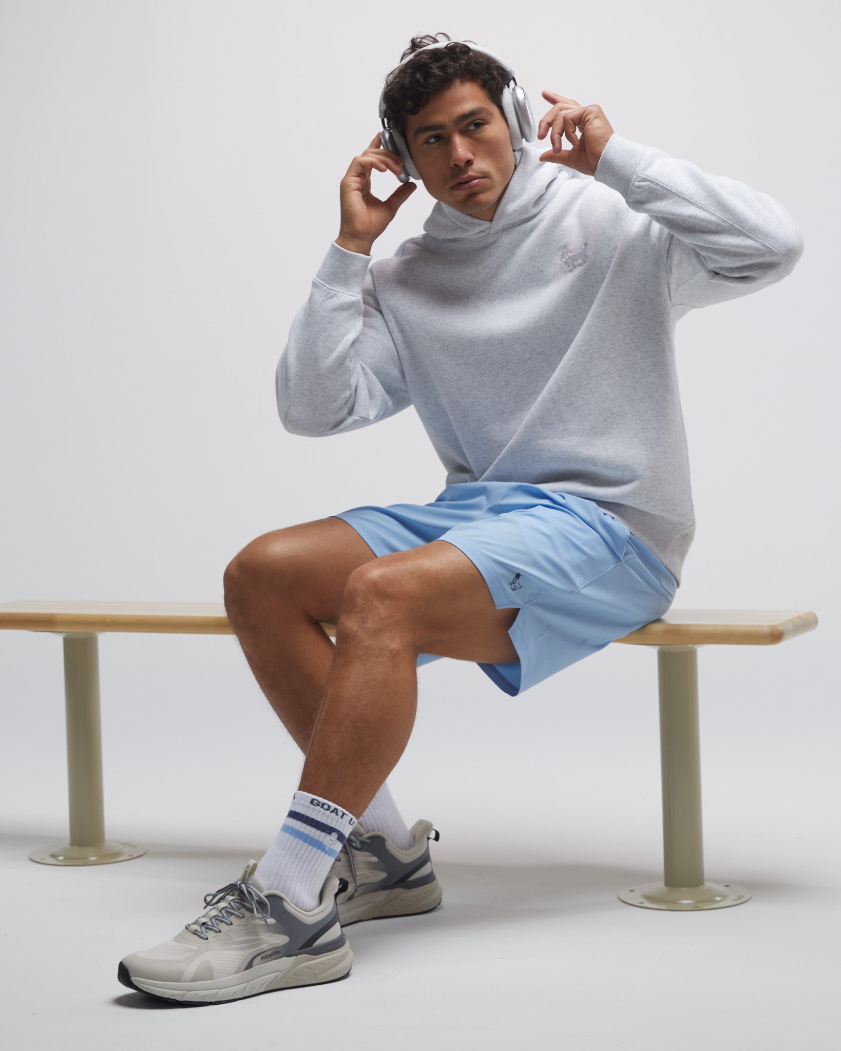 Man sitting on a bench wearing a light gray sweatshirt, blue shorts, and white sneakers.#color_carolina-blue