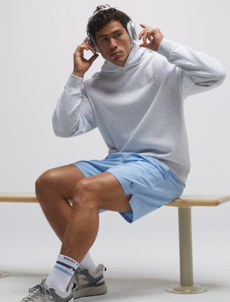 Man sitting on a bench wearing a light gray sweatshirt, blue shorts, and white sneakers.#color_carolina-blue