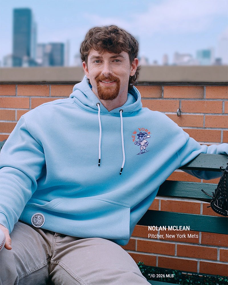 A man wearing a light blue pullover hoodie with a New York Mets graphic on the chest sits on a green bench against a brick wall and city skyline background. Text on the image identifies him as Nolan McLean, pitcher, New York Mets.