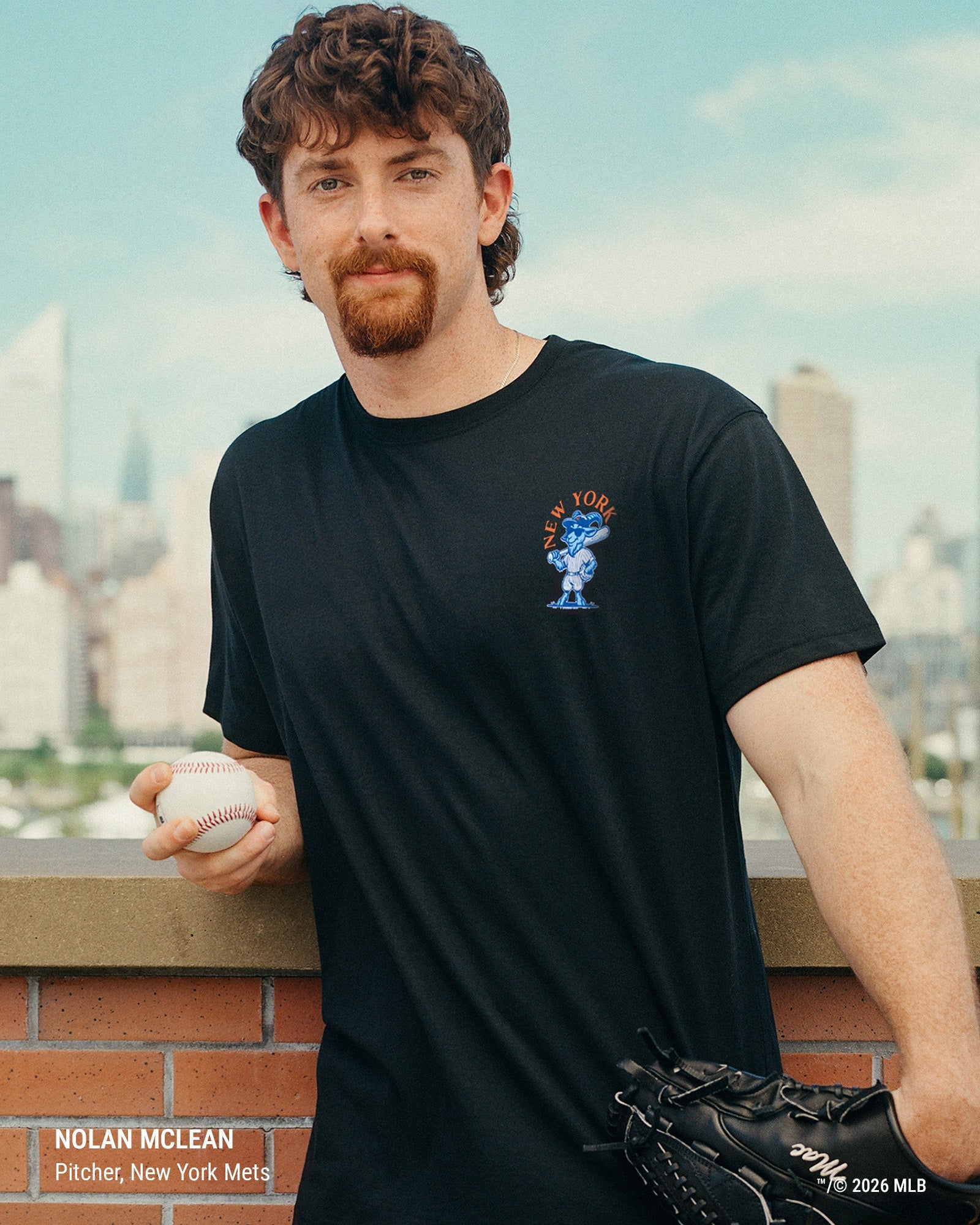 A man wearing a black short-sleeve t-shirt with a small blue and orange New York Mets graphic on the left chest, holding a baseball and wearing a black baseball glove. The background shows a blurred city skyline and sky.