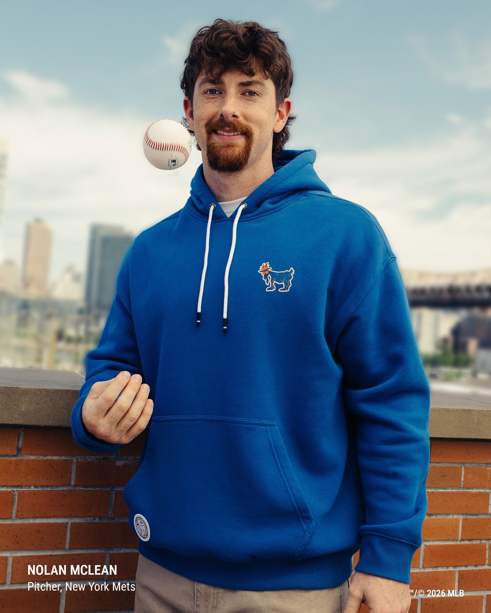A man wearing a royal blue pullover hoodie with white drawstrings and a small New York Mets dog logo on the left chest, posed outdoors against a city skyline with a baseball floating near his face.