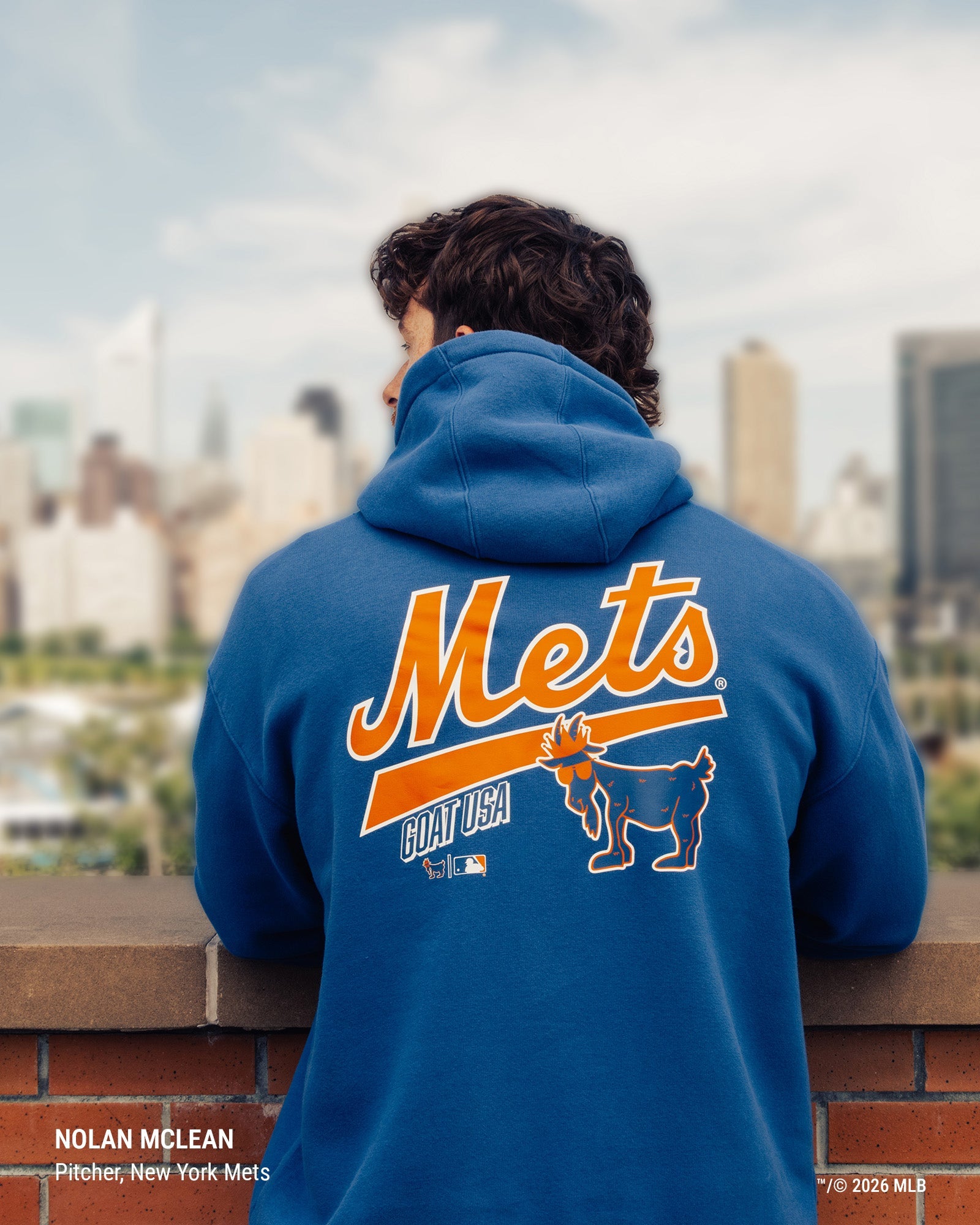 A man seen from behind wearing a royal blue hoodie with a large orange and white New York Mets graphic on the back, standing outdoors with a blurred city skyline in the background.